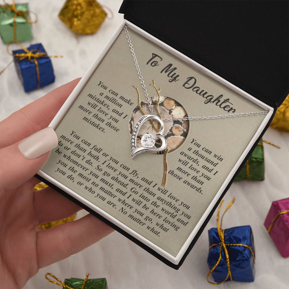 A hand holds a jewelry box containing the Daughter-A Thousand Awards - Forever Love Necklace in gold finish, showcasing a heart-shaped pendant with an inspirational message for "To My Daughter." In the background, there are small, colorful gift boxes.