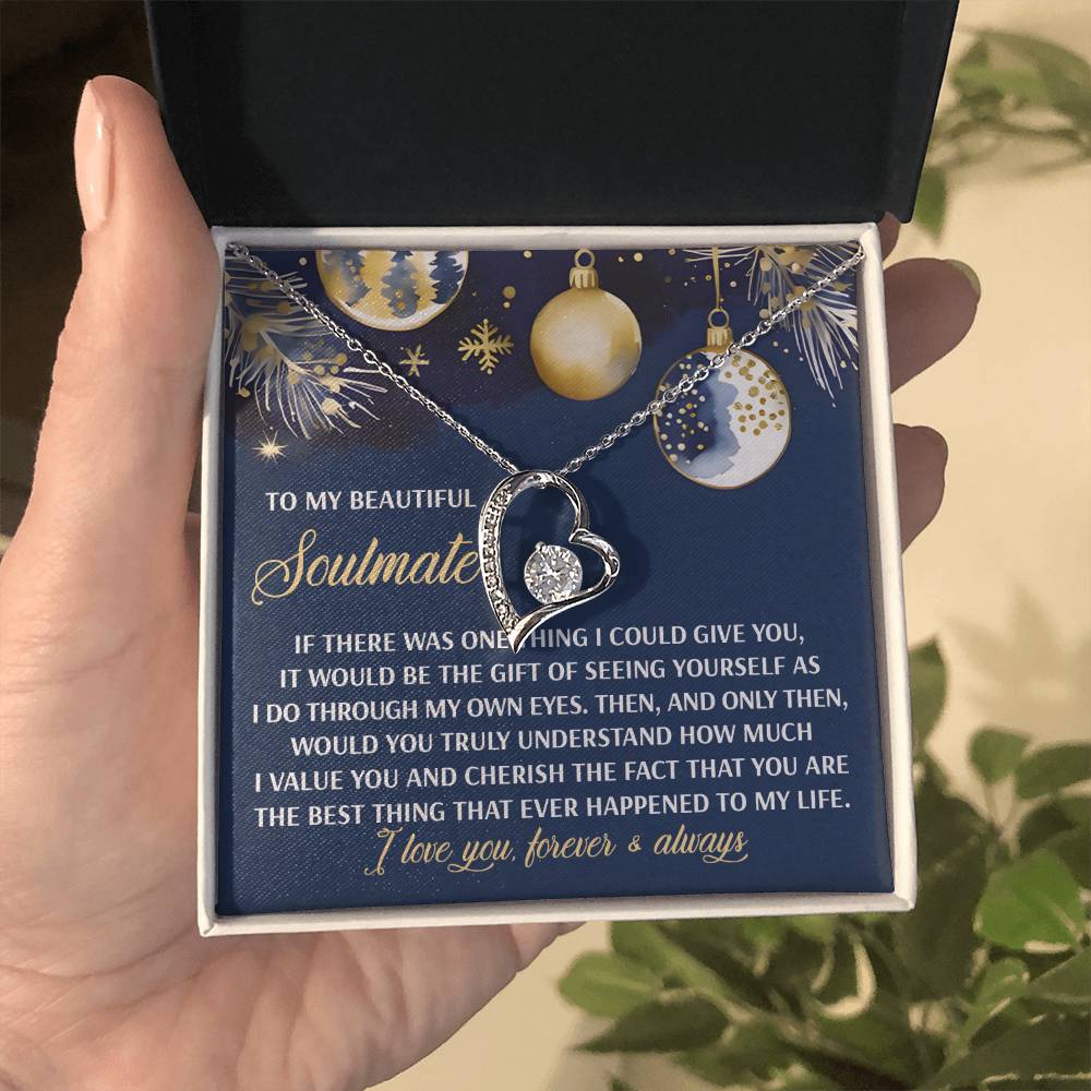 A hand holds an open box containing the Soulmate-One Thing - Forever Love Necklace, featuring a heart-shaped pendant adorned with a dazzling CZ crystal on a card with a message to a soulmate, all elegantly finished in white gold. The scene is beautifully decorated with gold and blue ornaments.