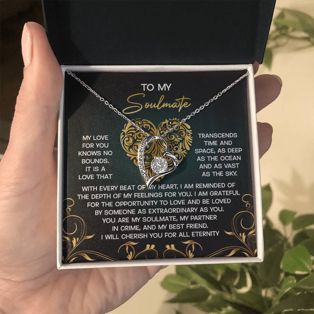 A hand holds the Soulmate-The Sky - Forever Love Necklace, featuring intertwined heart pendants in a box. The gold finish sparkles, accentuating CZ crystal accents. Inside, a decorative message about eternal love for a soulmate is displayed beautifully.