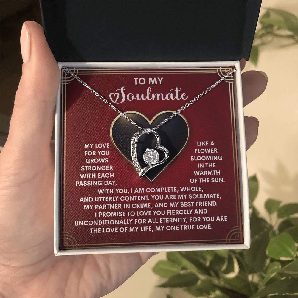 A hand presents an open gift box with the Soulmate-One True Love - Forever Love Necklace, a heart-shaped piece embellished with cubic zirconia stones. The lid displays sentimental text for a soulmate, surrounded by decorative heart motifs.