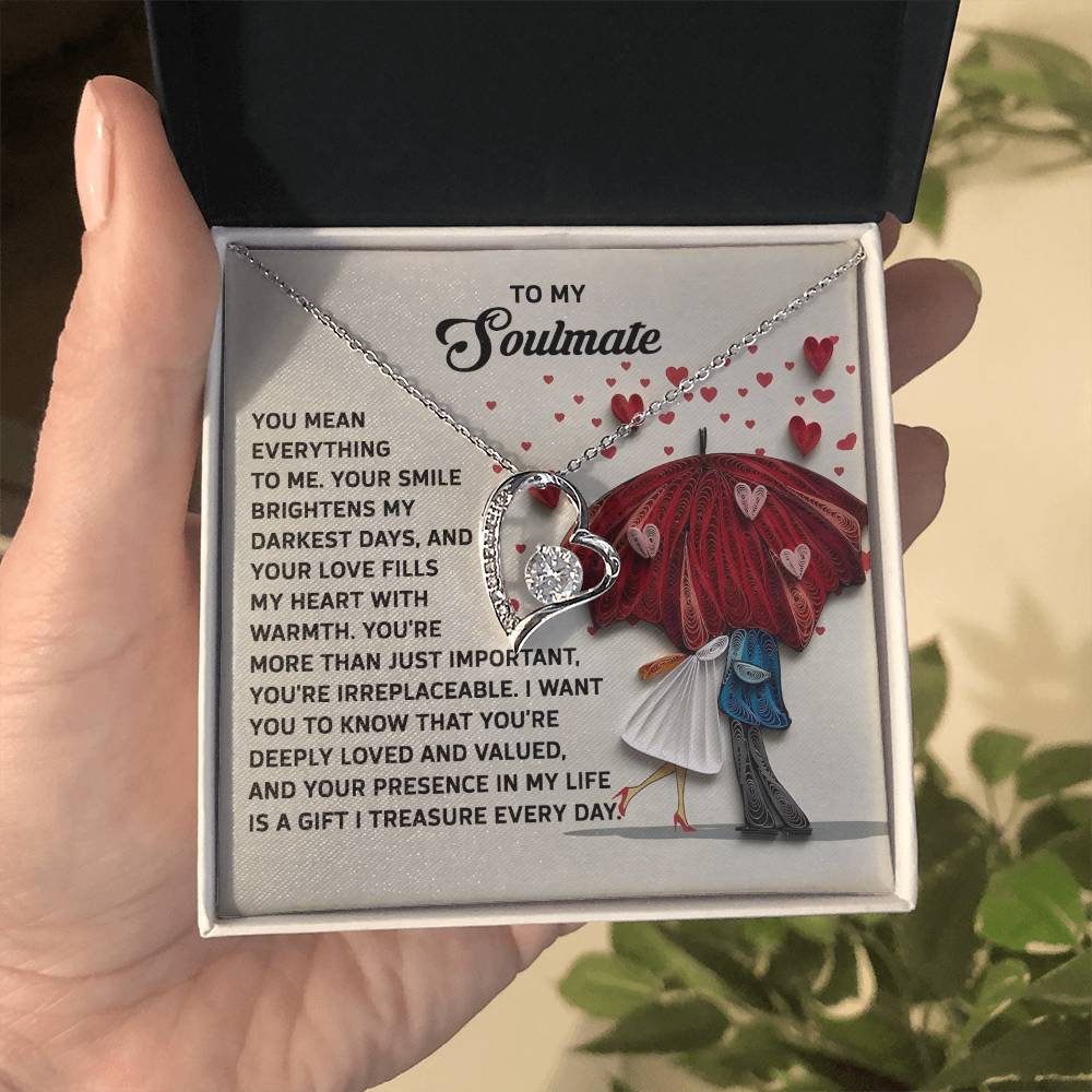 A hand holds an open box revealing a Forever Love Necklace, featuring a heart pendant with a white gold finish. In the background, an illustration of a couple under an umbrella and a printed romantic message enhances its charm. Product: Soulmate-Deeply Loved - Forever Love Necklace.