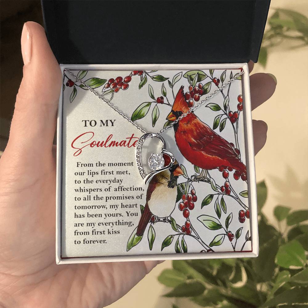 A hand holds an open jewelry box showcasing the Soulmate-First Kiss - Forever Love Necklace, adorned with a heart-shaped pendant in a dazzling gold finish. Alongside lies a card featuring text and two birds, completing this elegant presentation.