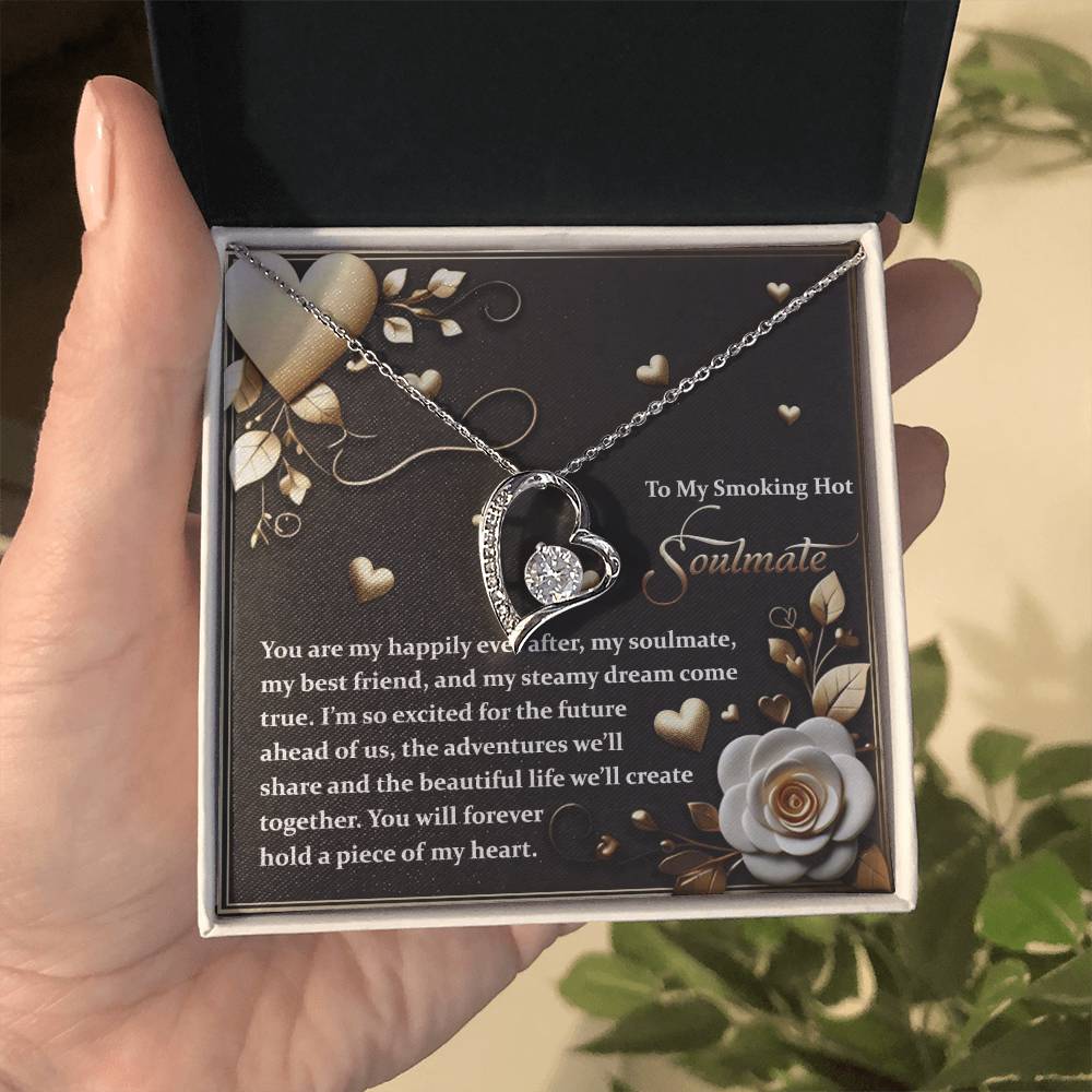 A hand holds an open box with the Soulmate-The Future Forever Love Necklace, showcasing a heart charm with interlocking hearts. With a gold finish and sparkling cubic zirconia, it rests beneath the message "To My Smoking Hot Soulmate" against a floral backdrop.