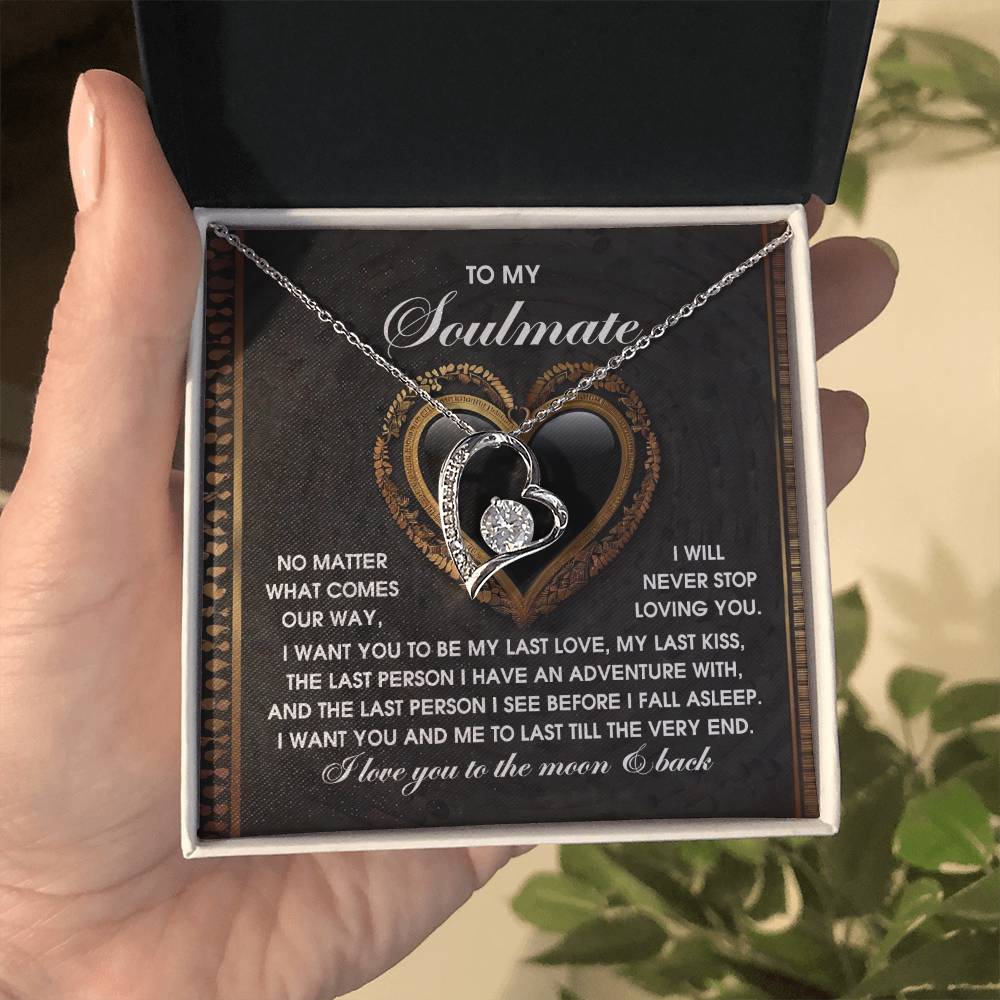 A hand holds the Soulmate-The Very End - Forever Love Necklace featuring intertwined hearts and a dazzling cubic zirconia stone, inside a box with a romantic message about love and soulmates, enhancing its charm with a 14k white gold finish.