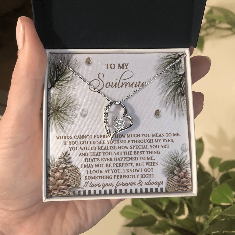 A hand holds the Soulmate-Perfectly Right - Forever Love Necklace, showcasing its heart pendant adorned with a sparkling CZ crystal. Elegantly presented in a gift box, the card features a romantic message and is decorated with pine cones and branches, enhancing the necklace's luxurious gold finish.