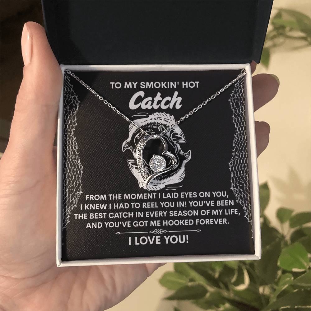 A hand displays an open box revealing the Soulmate-Got Me Hooked - Forever Love Necklace, featuring two fish encircling a heart with sparkling CZ crystal, accompanied by the message, "To my smokin' hot catch... I love you!" A plant decorates the background.