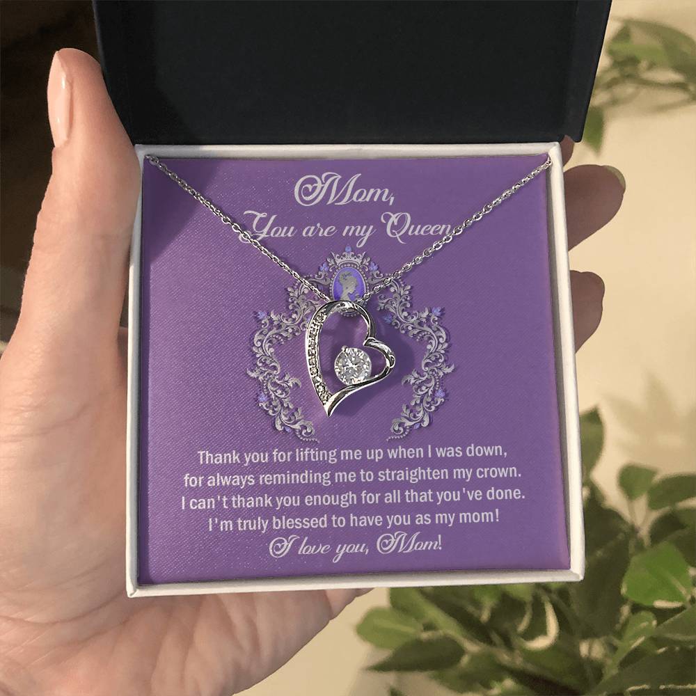 A hand holds an open box revealing a heart-shaped cubic zirconia pendant necklace against a purple background. The box's lid contains a heartfelt message addressed to "Mom, You are My Queen." This "To Mom, Belongs To Me - Forever Love Necklace" makes the perfect personalized gift for any occasion.