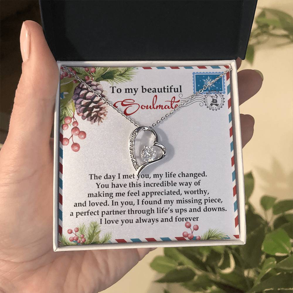 A hand holds a jewelry box containing the Soulmate-Perfect Partner - Forever Love Necklace, elegantly finished in gold. Inside, a heartfelt message expresses love and appreciation to a soulmate, framed by a decorative border adorned with pinecones and foliage.