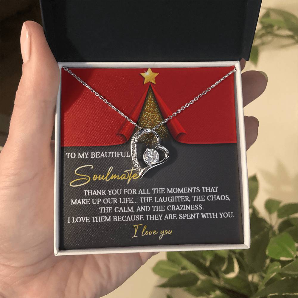 A hand presents an open gift box revealing the Soulmate-The Moments - Forever Love Necklace, a heart-shaped keepsake adorned with a CZ crystal and a white gold finish. The cover of the box displays an appreciation message for a soulmate, enhanced with an elegant red and gold design.