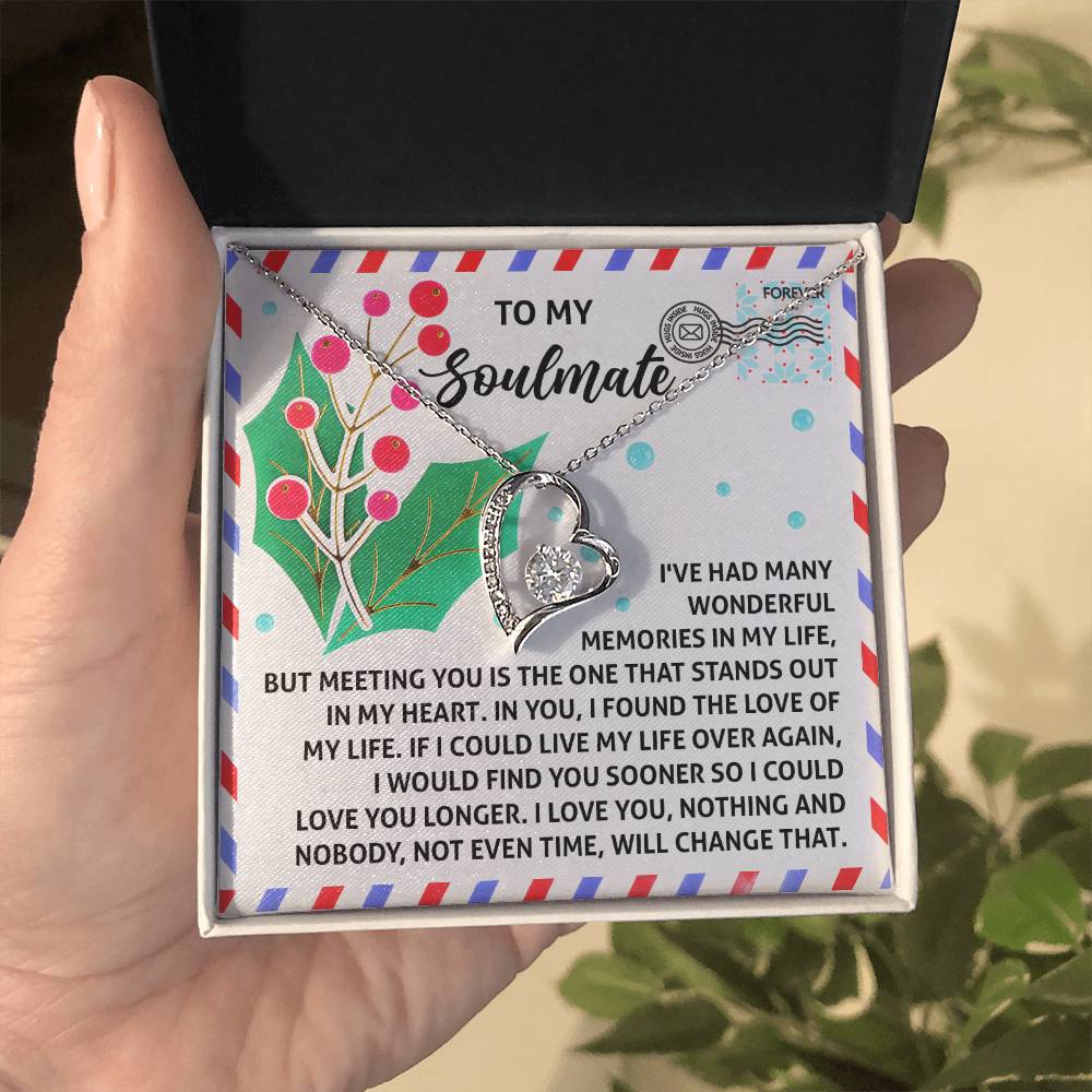 A hand holds an open box, unveiling the Soulmate-Love You Longer - Forever Love Necklace with a white gold finish. The heart-shaped pendant, embellished with sparkling CZ crystals, rests inside the box adorned with a message to a soulmate against a decorative background of holly and colorful dots.