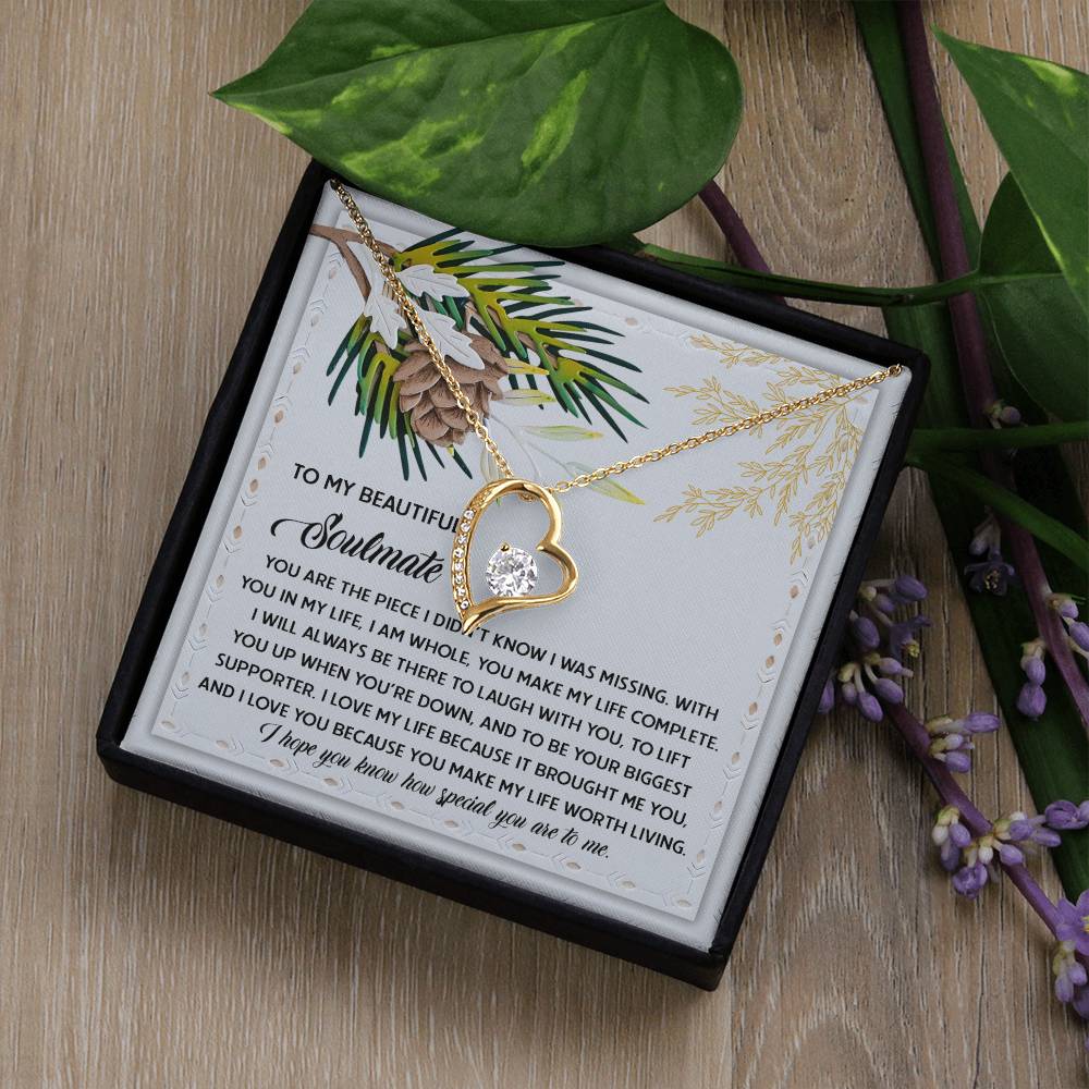 The Soulmate-Biggest Supporter - Forever Love Necklace, featuring a gold heart shape with a cubic zirconia stone, rests elegantly on a decorative card with a sentimental message. Encased in a black box adorned with green leaves and purple flowers, it boasts a timeless white gold finish.