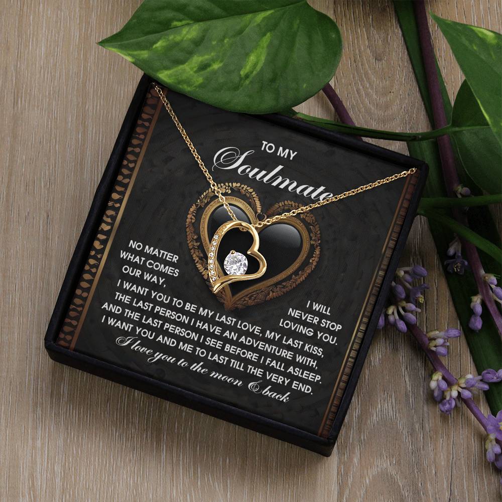 The Soulmate-The Very End - Forever Love Necklace features a gold heart-shaped design with a sparkling diamond, crafted in 14k white gold finish, and sits elegantly on a box with a romantic message, framed by lush green leaves and vibrant purple flowers.