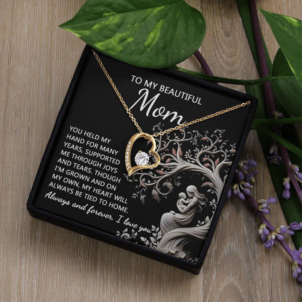 The "To Mom, On My Own - Forever Love Necklace," featuring a gold heart-shaped pendant with an inset Cubic Zirconia stone and accompanied by a card reading "To my beautiful mom," is beautifully displayed on a wooden surface adorned with green leaves and purple flowers, encapsulating the essence of the perfect personalized gift.