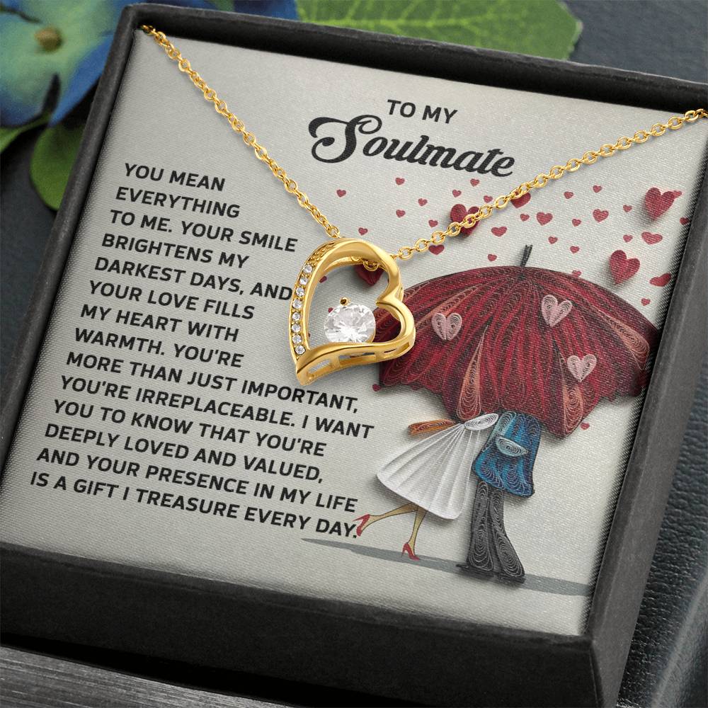 A gold heart necklace with CZ crystal, named Soulmate-Deeply Loved - Forever Love Necklace, rests on a box featuring romantic text and a couple under a red umbrella.