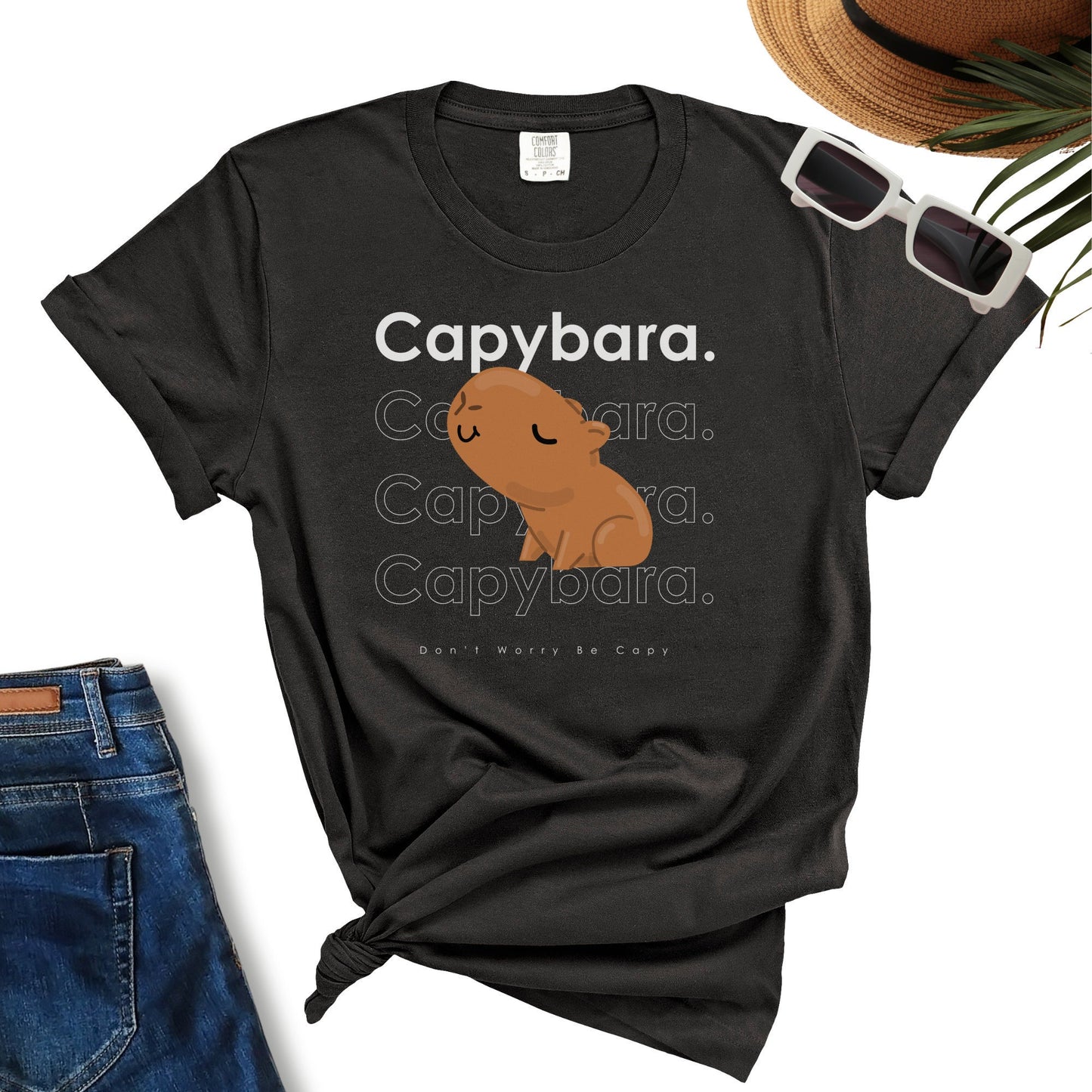 Black “Don’t Worry Be Capy Cute” T-shirt featuring a charming capybara graphic and the text "Capybara" repeated in shades of grey and white. Ideal for any Capybara Lover T-Shirt collection, it pairs perfectly with jeans, sunglasses, and a hat placed beside it.