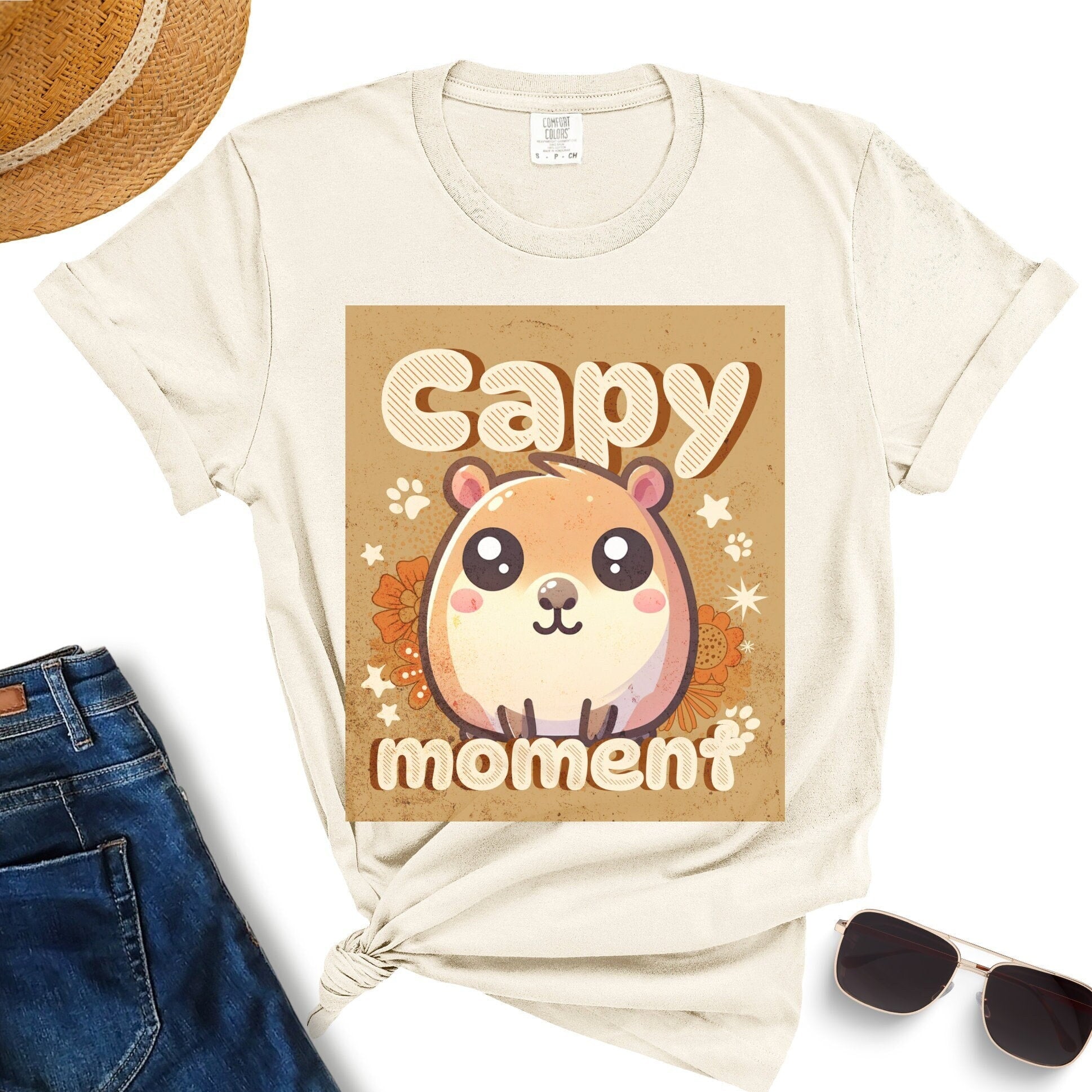 Beige Capy Moment Cute Capybara T-Shirt, perfect for both women and men, showcasing a kawaii capybara with the text "Capy moment." This adorable crewneck tee makes an ideal gift for capybara lovers, besties, girlfriends, boyfriends, and moms. It's styled here with a straw hat, sunglasses, and jeans.