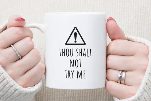 Hands holding a white "Thou Shalt Not Try Me Coffee Mug for Moms & Teachers - Quirky Gift Idea" with a caution symbol above it— the perfect gift for moms and teachers who appreciate religious humor.