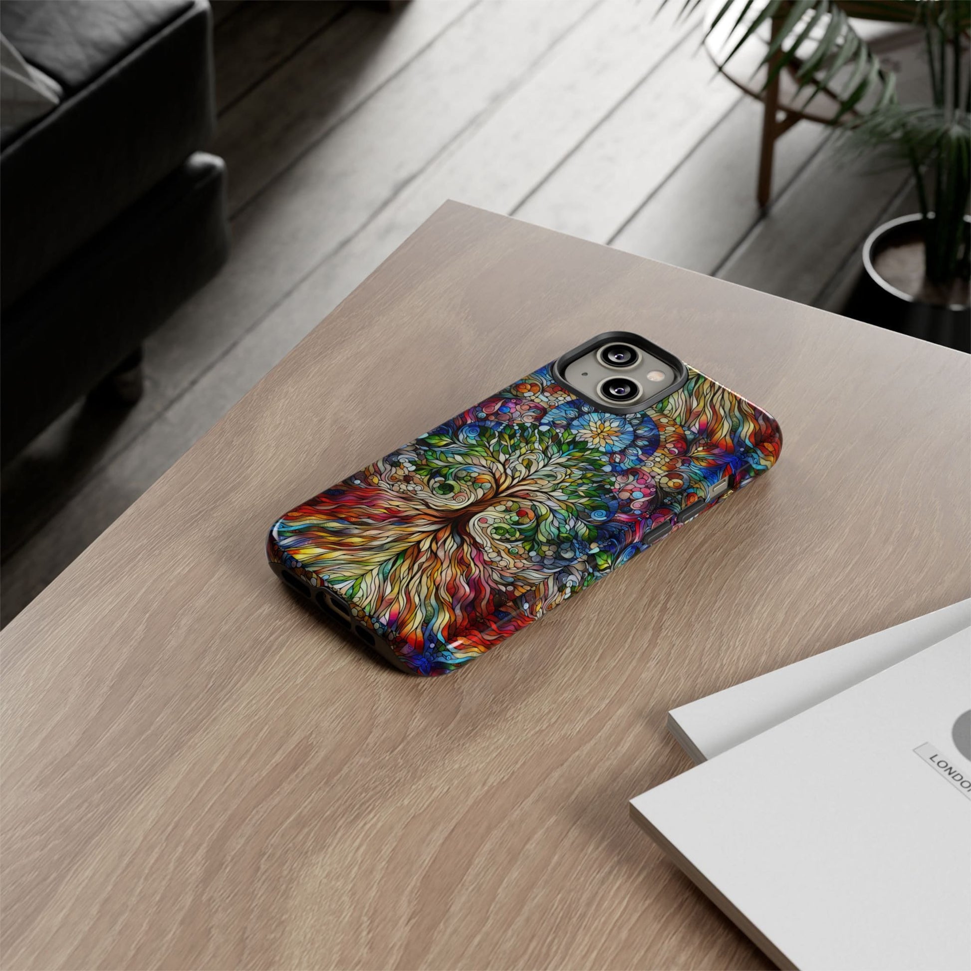 A smartphone adorned with the Celtic Tree of Life Aesthetic Phone Case is placed on a wooden table next to a booklet and a potted plant.