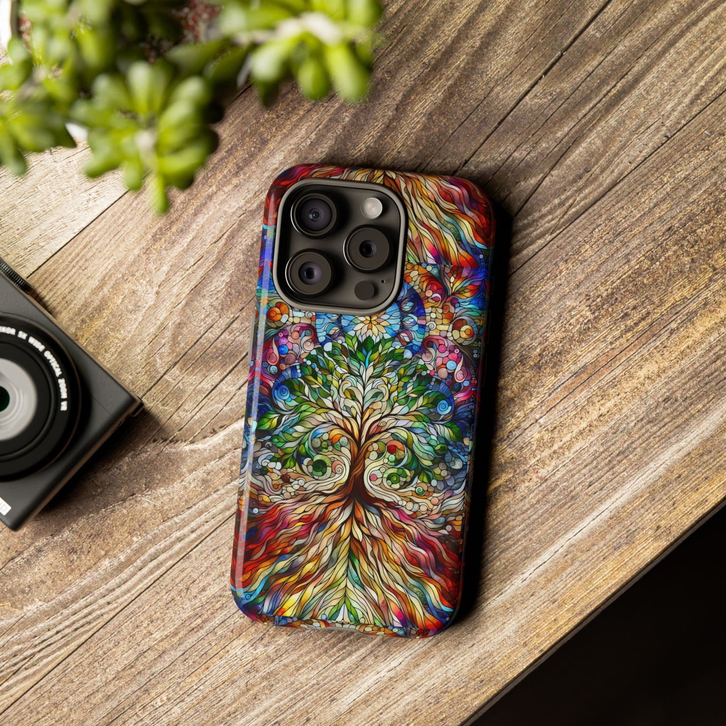 The Celtic Tree of Life Aesthetic Phone Case, featuring an ornate and colorful design, rests on a wooden surface alongside a small camera and a green plant.