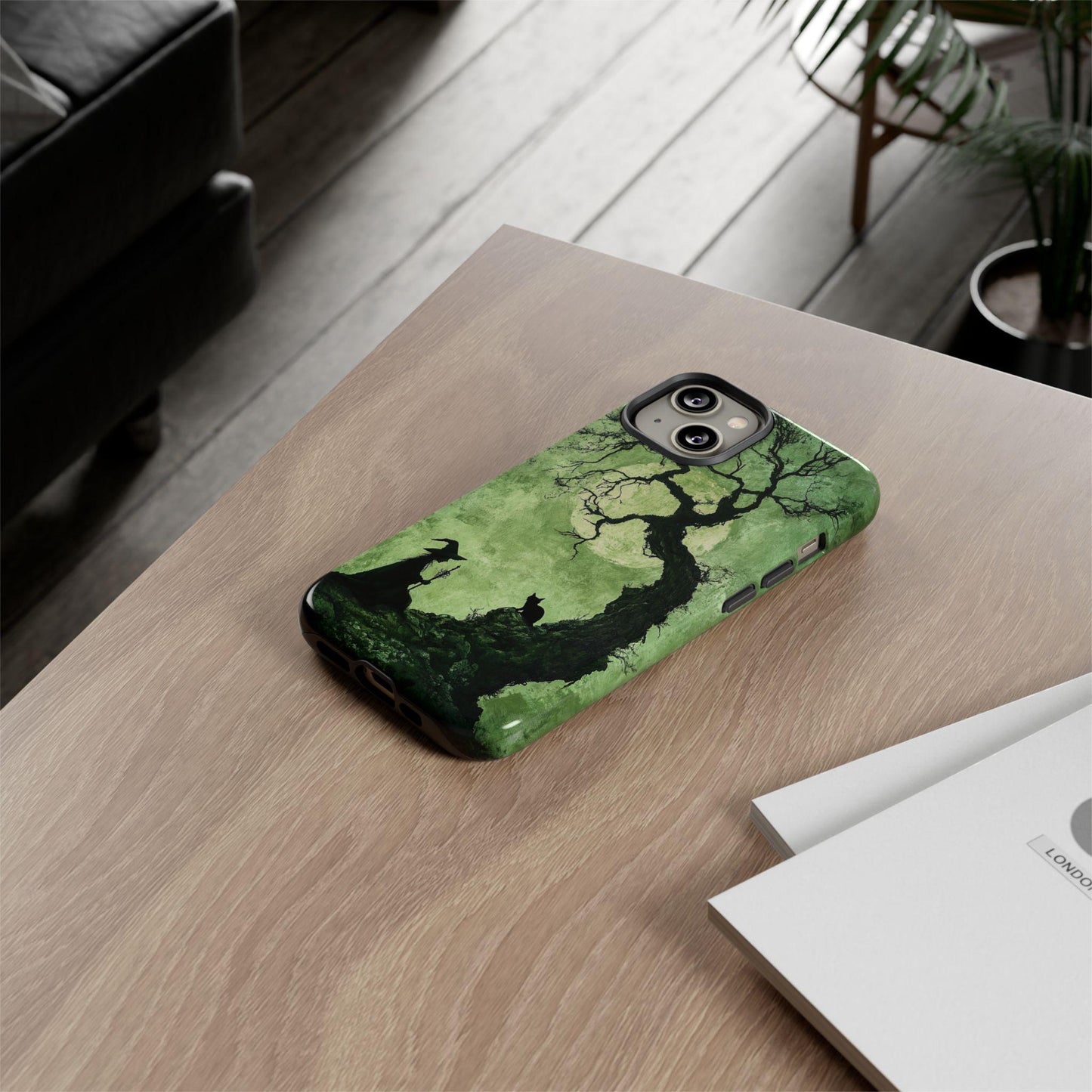 A wooden table holds a smartphone encased in a Spooky Witch With Black Cat Phone Case, accompanied by a nearby notebook and surrounded by a plant and chair, creating an ideal scene for those who adore black cats.