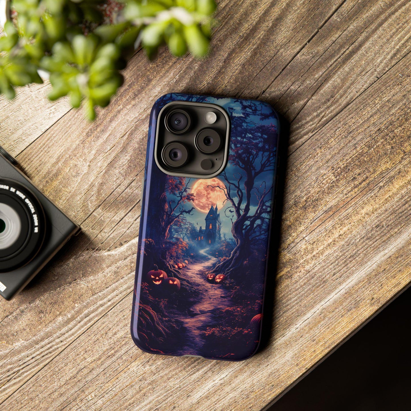 An iPhone with a Haunted Mansion Phone Case, featuring spooky bats and pumpkins, lays on a wooden surface beside a camera, enhancing the eerie charm.