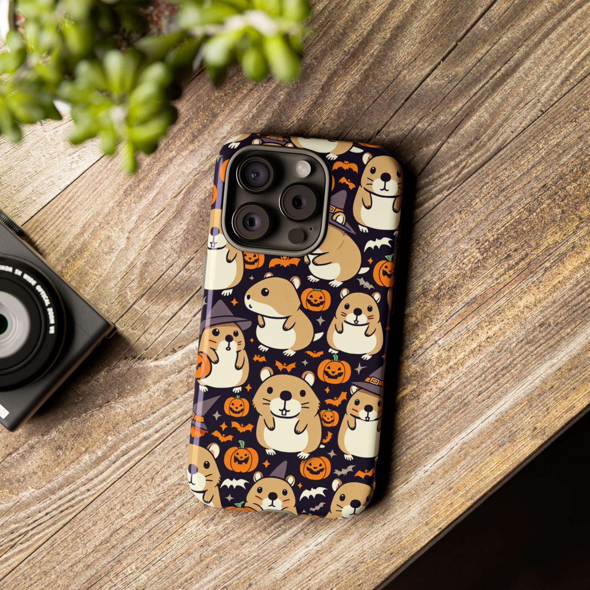 A smartphone equipped with a Halloween Cute Hamster Phone Case, featuring adorable cartoon dogs, pumpkins, and bats, sits on a wooden surface next to a small plant and a camera. This charming accessory is ideal for any hamster lover's gift collection.