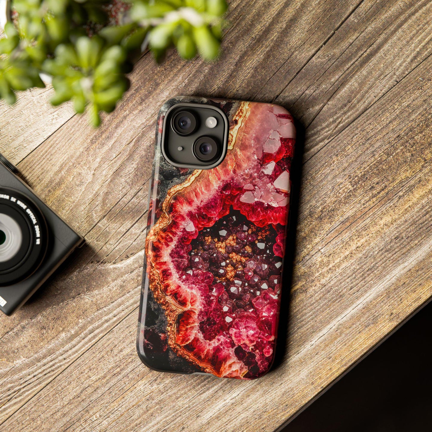 A smartphone featuring the January Birthstone Garnet Crystal Geode Design Phone Case lies on a wooden surface beside a camera and a small green plant, highlighting its exquisite geode pattern that embodies elegance.