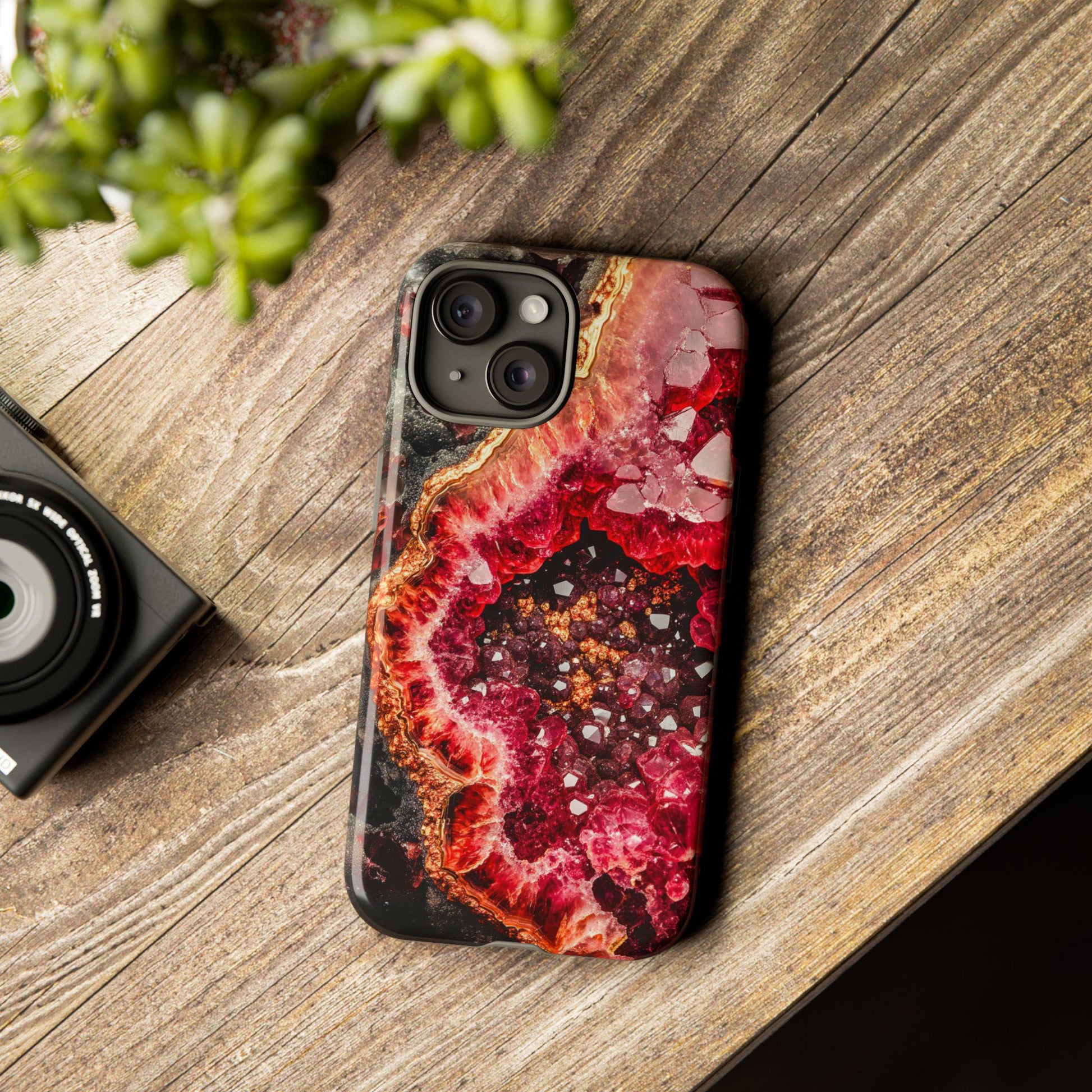 A smartphone featuring the January Birthstone Garnet Crystal Geode Design Phone Case lies on a wooden surface beside a camera and a small green plant, highlighting its exquisite geode pattern that embodies elegance.