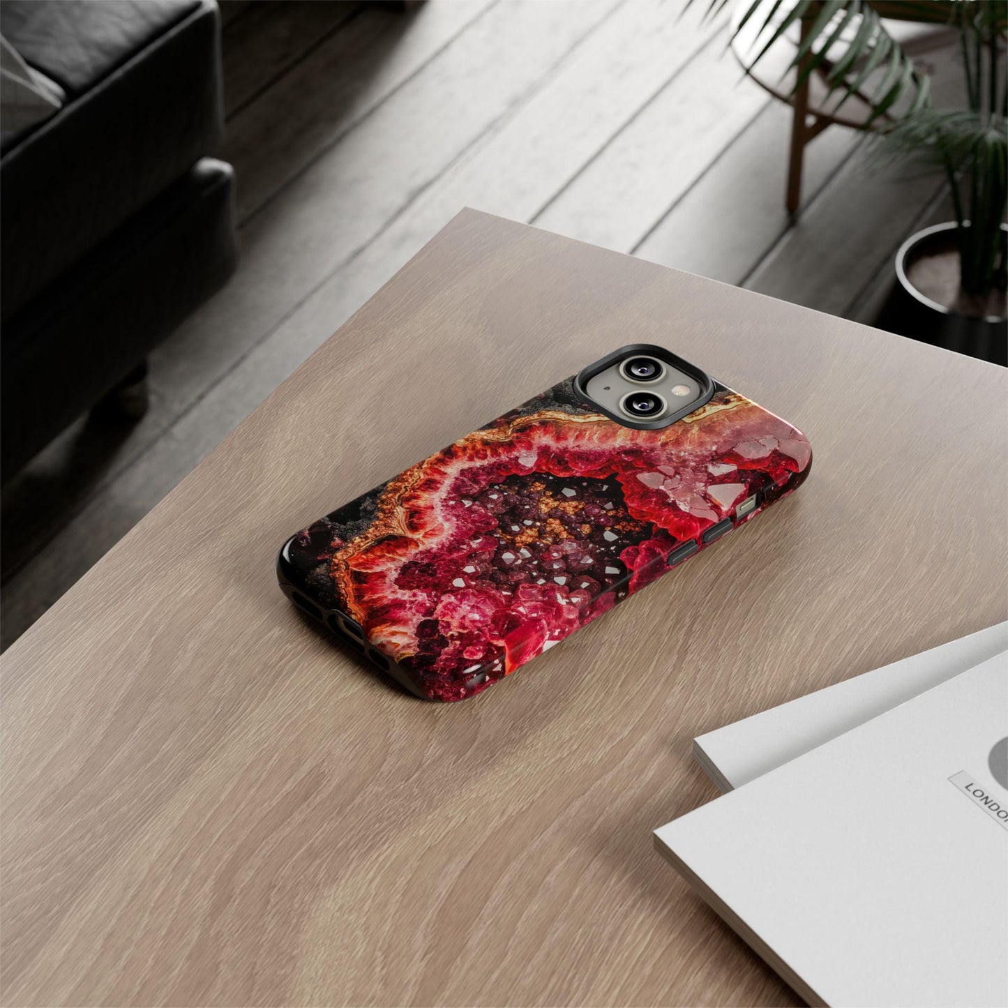 The January Birthstone Garnet Crystal Geode Design Phone Case, perfect for iPhone 15, 14, 13, Samsung, and Google Pixel users who love geodes and garnets, sits vibrantly on a wooden table near a stack of papers and a potted plant.