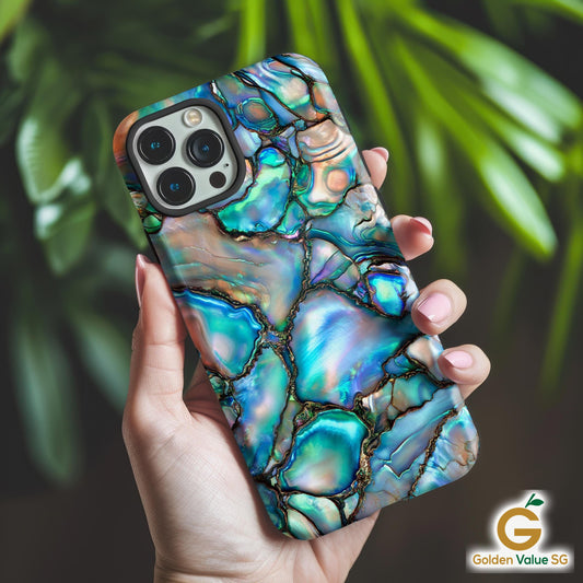 A lady&#39;s hand is holding an iphone Abalone Shell Design Phone Case featuring the iridescent beauty of the case delivers a mesmerizing play of colors that mimic the captivating allure of gemstones. Against the background of some green plants.