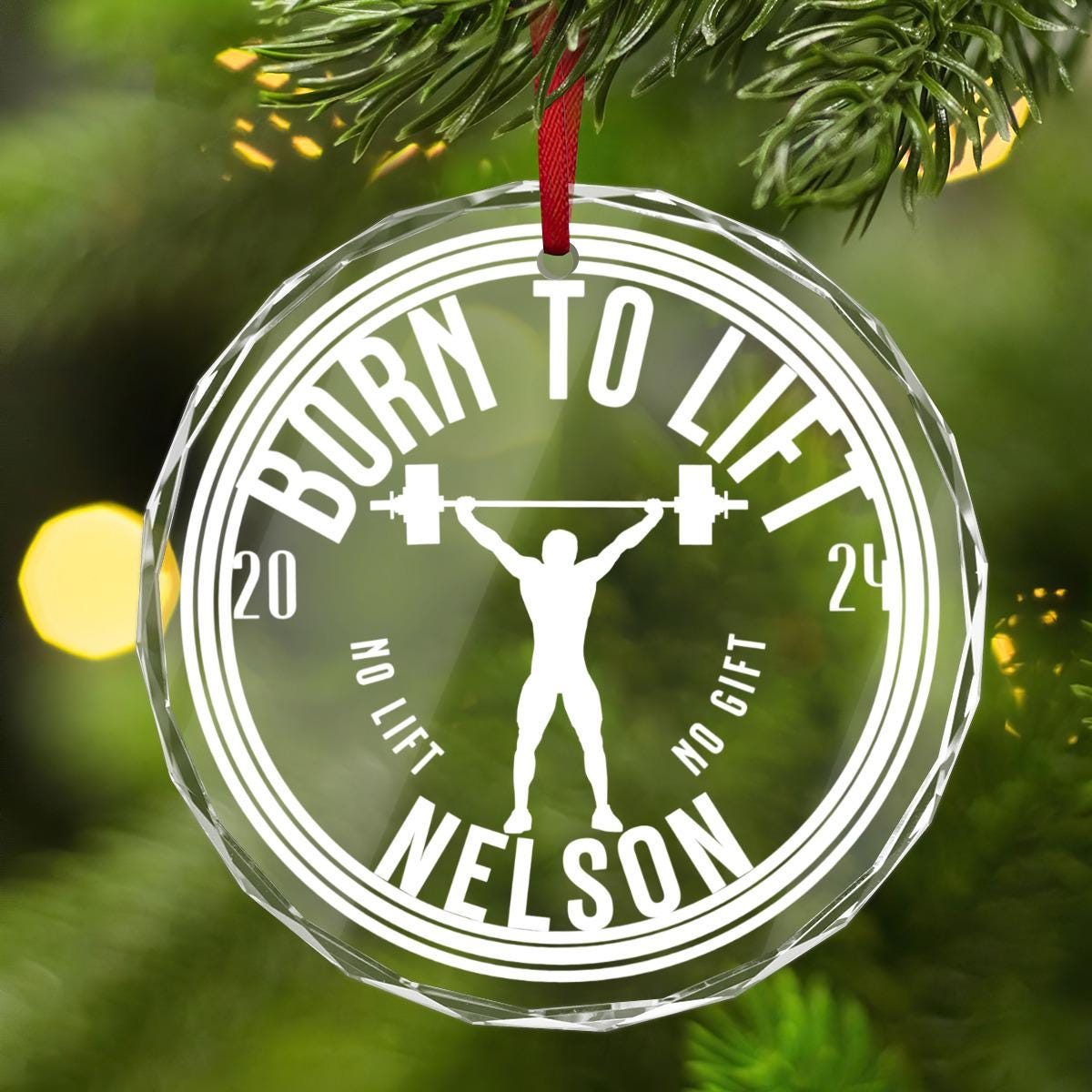 A Custom Weightlifting CRYSTAL GLASS Ornament hanging on a Christmas tree. 
It showcases a weightlifter&#39;s silhouette triumphantly holding a barbell overhead with the playful text &quot;BORN TO LIFT&quot; and &quot;NO LIFT, NO GIFT&quot; 2024 and the name Nelson.