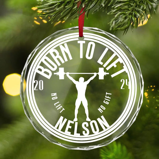 A Custom Weightlifting CRYSTAL GLASS Ornament hanging on a Christmas tree. 
It showcases a weightlifter&#39;s silhouette triumphantly holding a barbell overhead with the playful text &quot;BORN TO LIFT&quot; and &quot;NO LIFT, NO GIFT&quot; 2024 and the name Nelson.