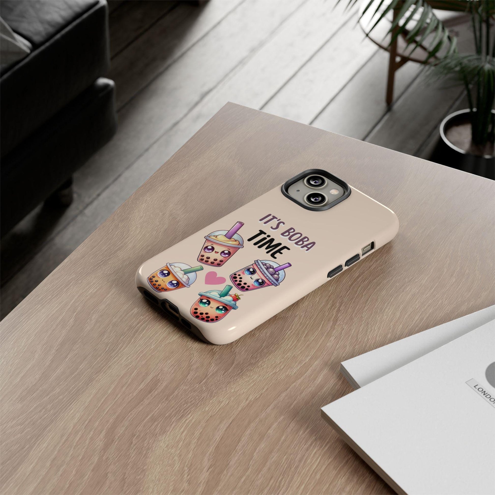 The Kawaii Boba Phone Case, featuring cartoon boba tea cups and "It's Boba Time" text, is perfect for iPhone 16 Pro Max and various Samsung Galaxy models. Ideal for boba lovers who cherish cute designs, it sits on a wooden surface beside some papers.