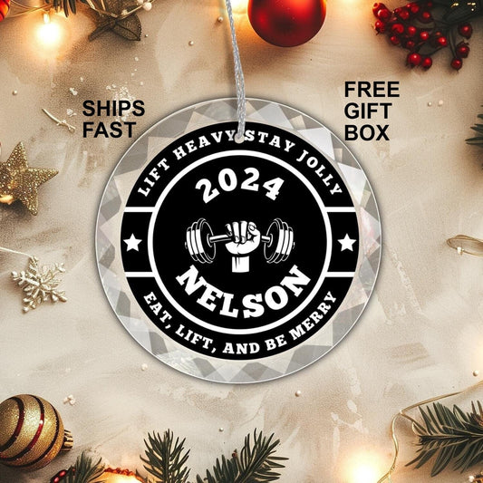 Golden Value SG&#39;s Custom Weightlifting GLASS Weight Plate Ornament features a hand holding a dumbbell, with the text &quot;2024 Nelson&quot; and the phrases &quot;Lift Heavy Stay Jolly&quot; and &quot;Eat, Lift, and Be Merry.