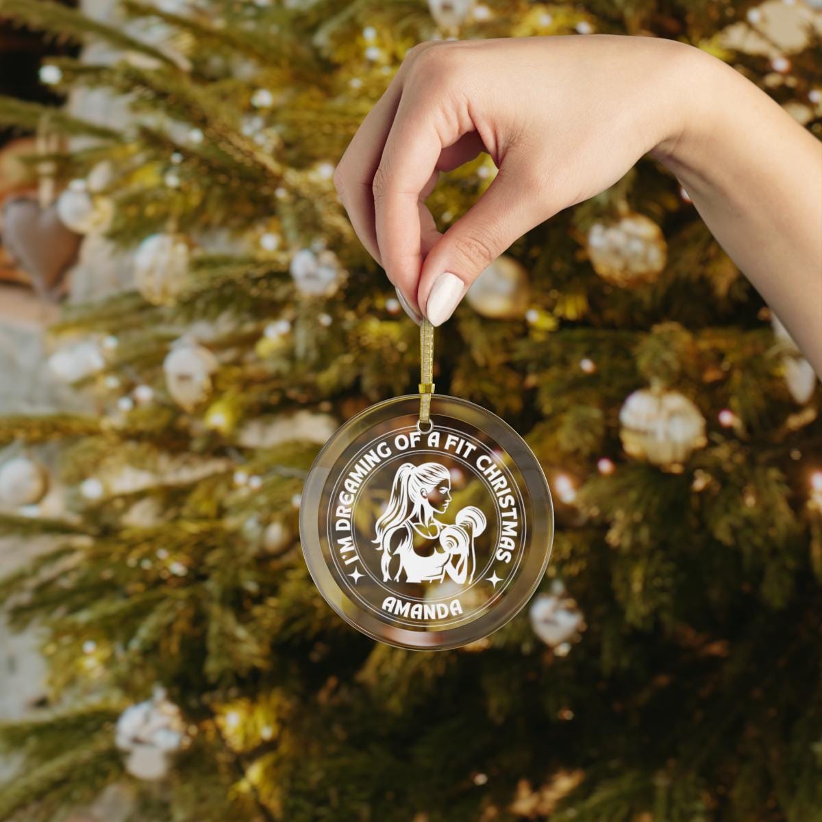 A hand holds a Personalized Weightlifting Glass Ornament featuring the custom text "I'm dreaming of a fit Christmas Amanda" in front of a decorated tree, making it an ideal gift for female weightlifters.