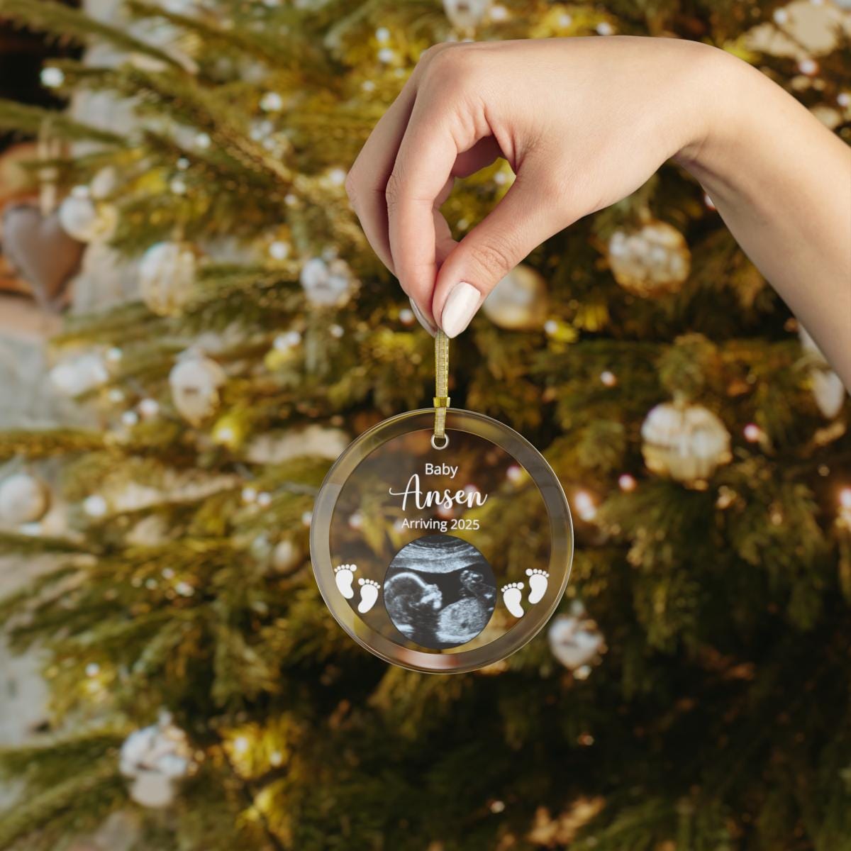 A hand holds a transparent Custom Ultrasound Photo GLASS Ornament, adorned with baby footprints and personalized with the text "Baby Anson Arriving 2025," set against the backdrop of a beautifully decorated Christmas tree.