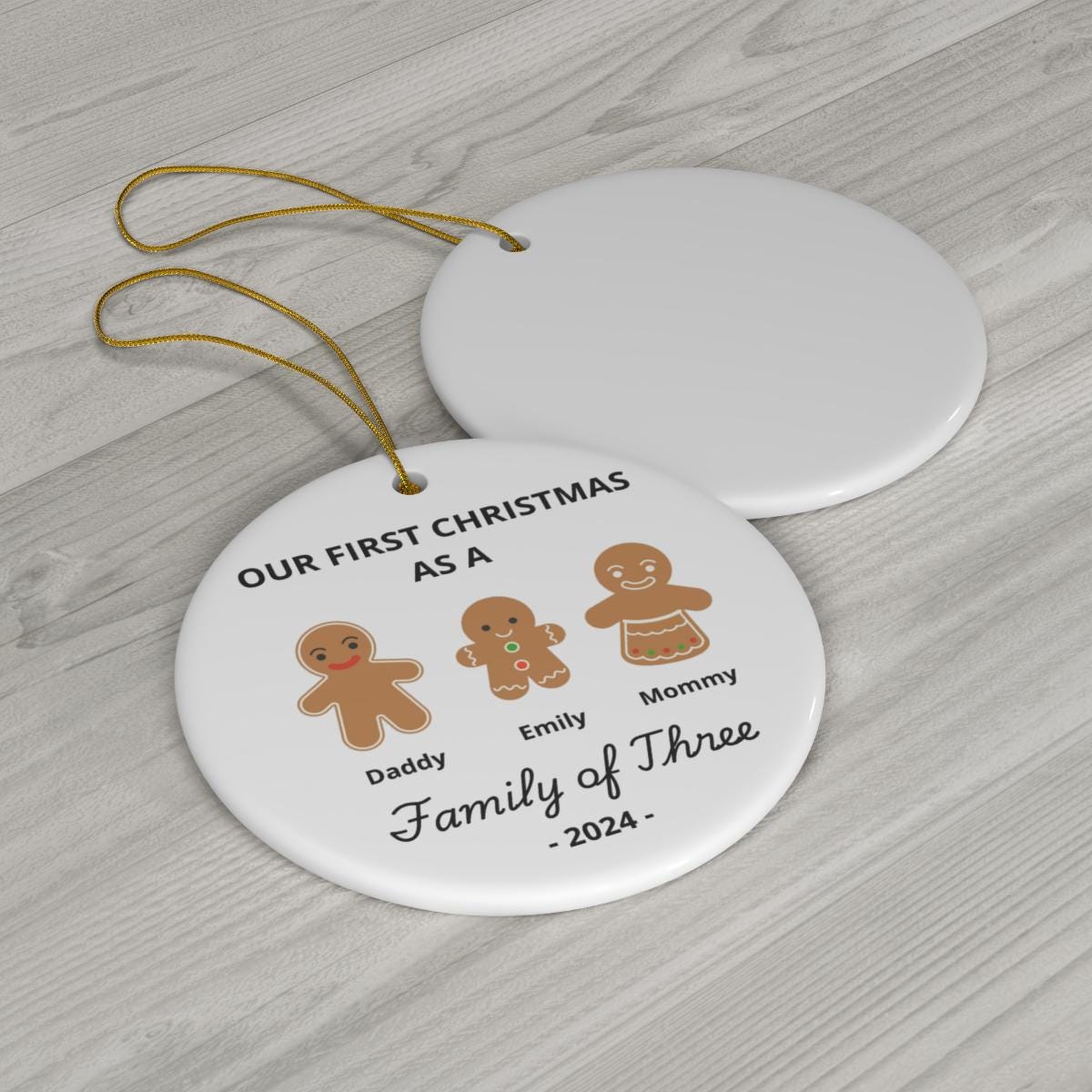 On a wooden surface, there are two circular ornaments. One is plain, while the other is a Personalized Family Of Three Christmas Ornament featuring gingerbread figures labeled with custom names "Daddy," "Emily," and "Mommy." It says, "Our First Christmas as a Family of Three - 2024," commemorating Baby's First Xmas.