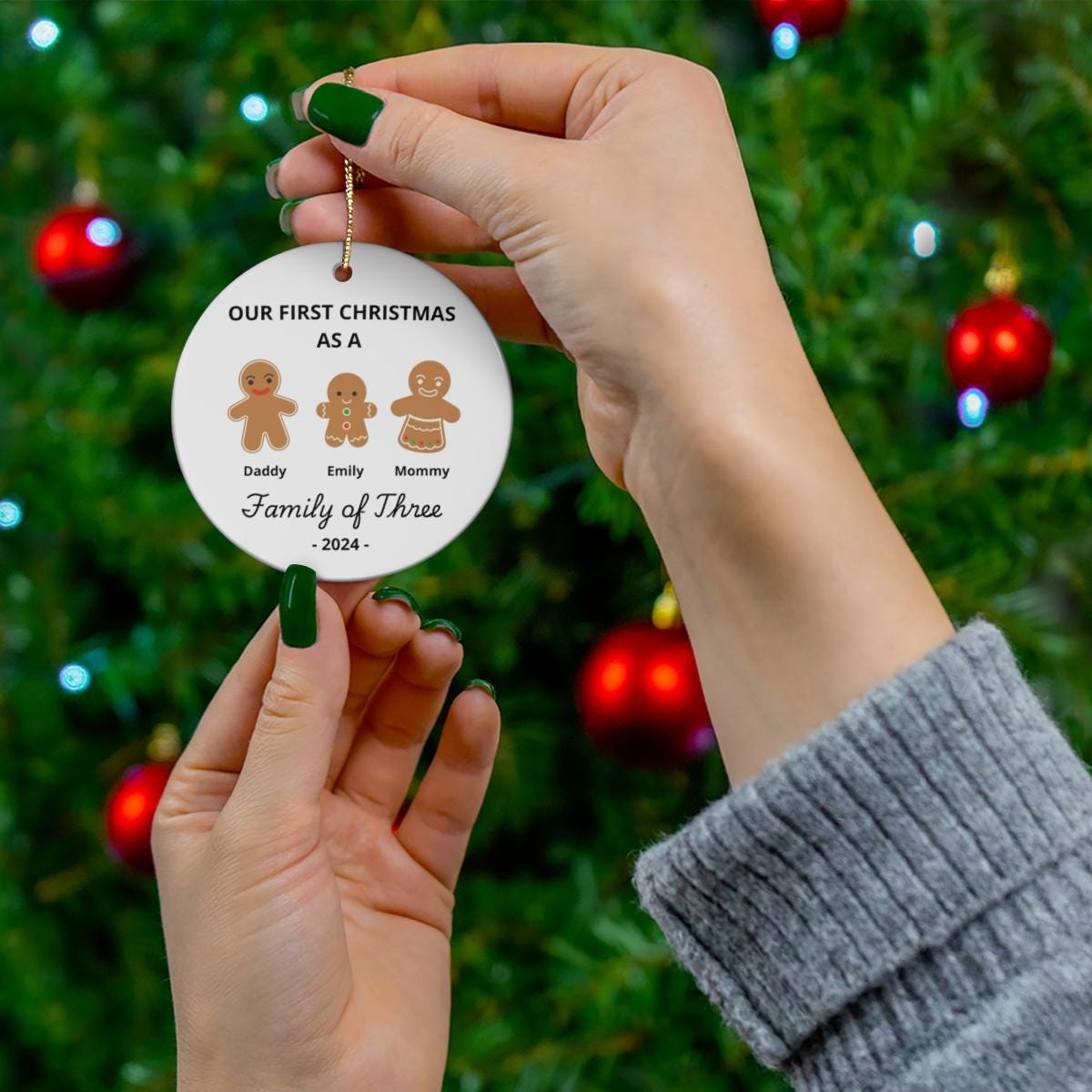 Hands hold a Personalized Family Of Three Christmas Ornament in front of a tree, featuring gingerbread figures and the year 2024. The ornament celebrates "Our First Christmas as a Family of Three," capturing the joy of Baby's First Xmas.