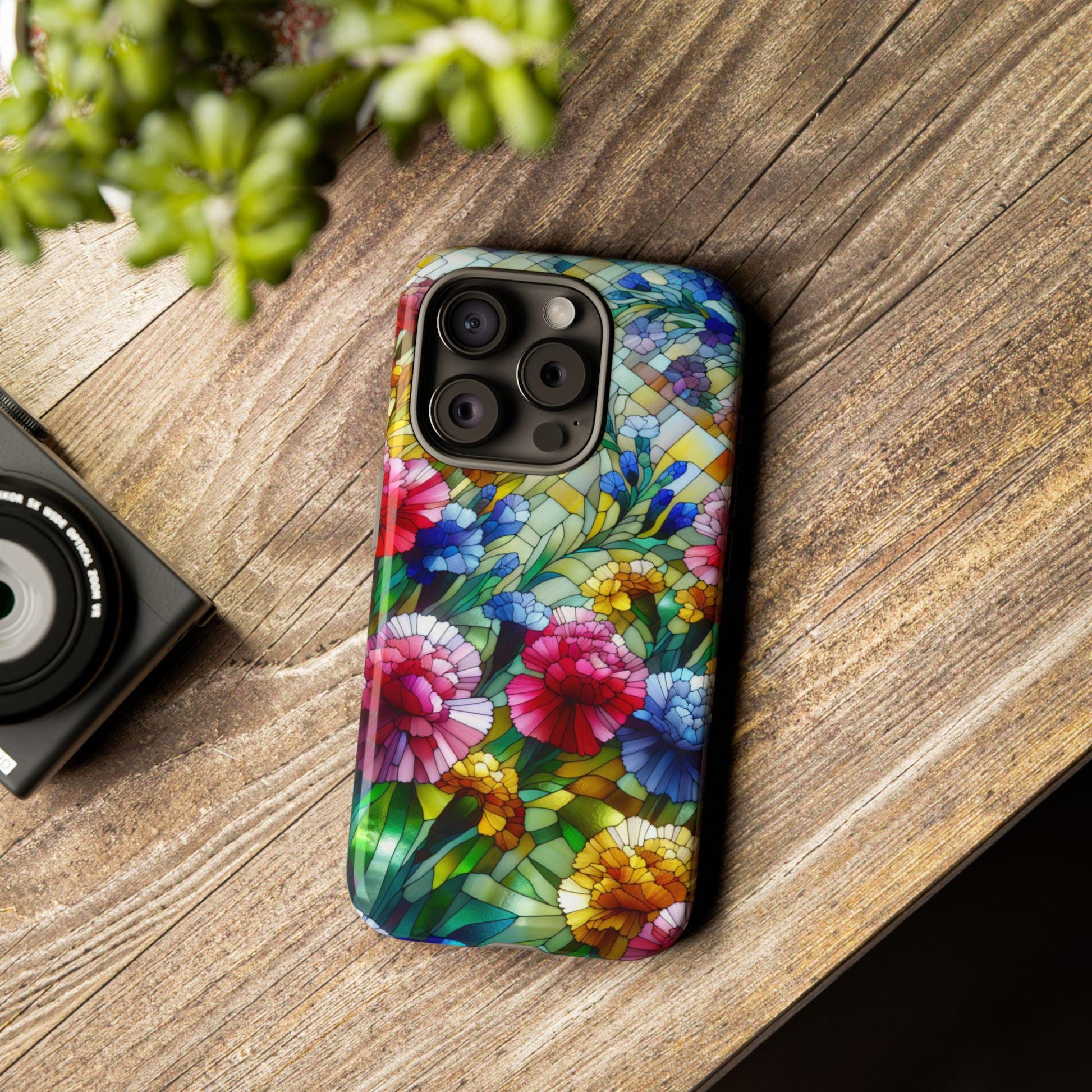 A smartphone with a January Birth Flower Carnation Phone Case Faux Stained Glass Cover sits on a wooden table, alongside a camera and a lush green plant.
