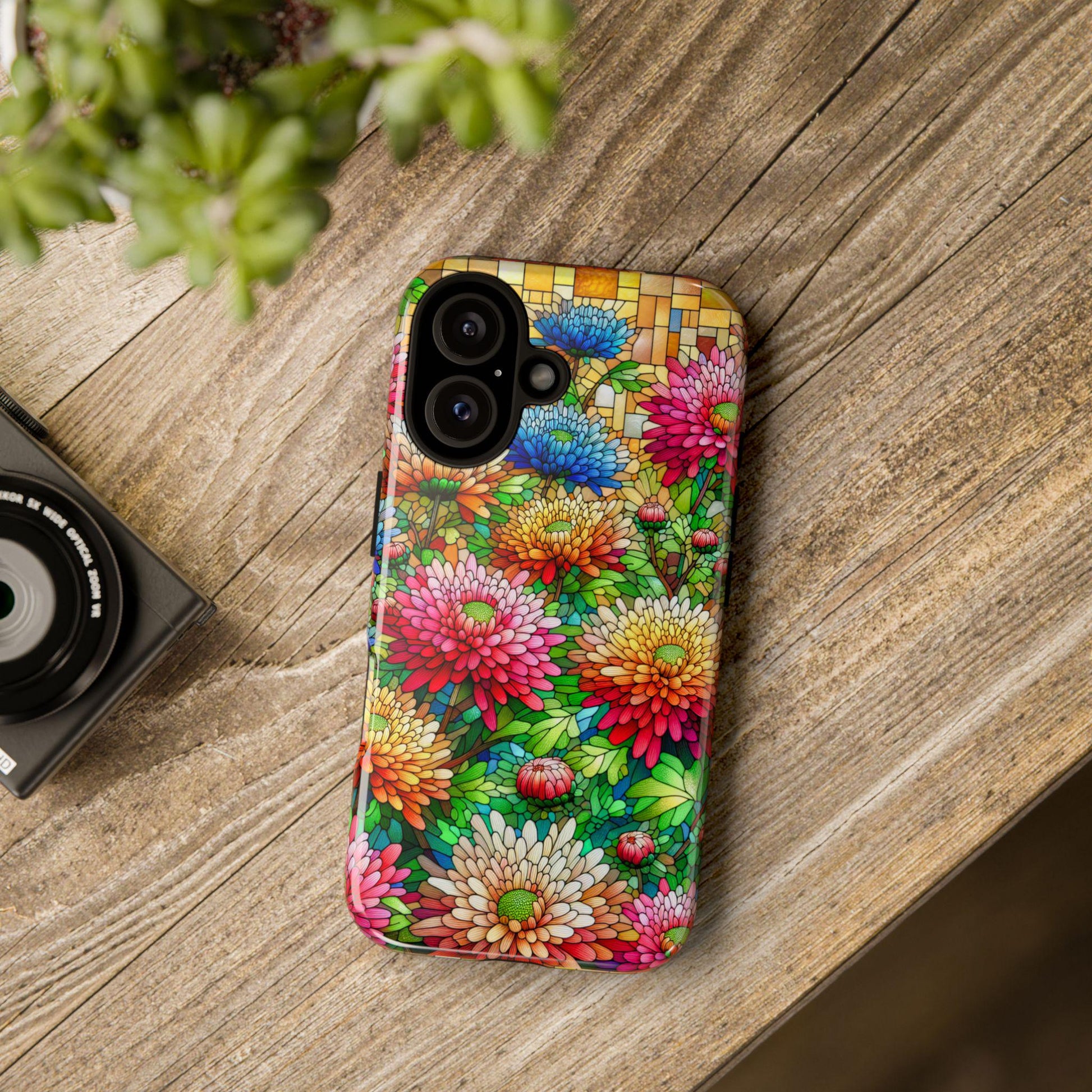 An iPhone with a November Birth Flower Chrysanthemum Faux Stained Glass Phone Case lies on a wooden surface beside a small black camera and a green plant.