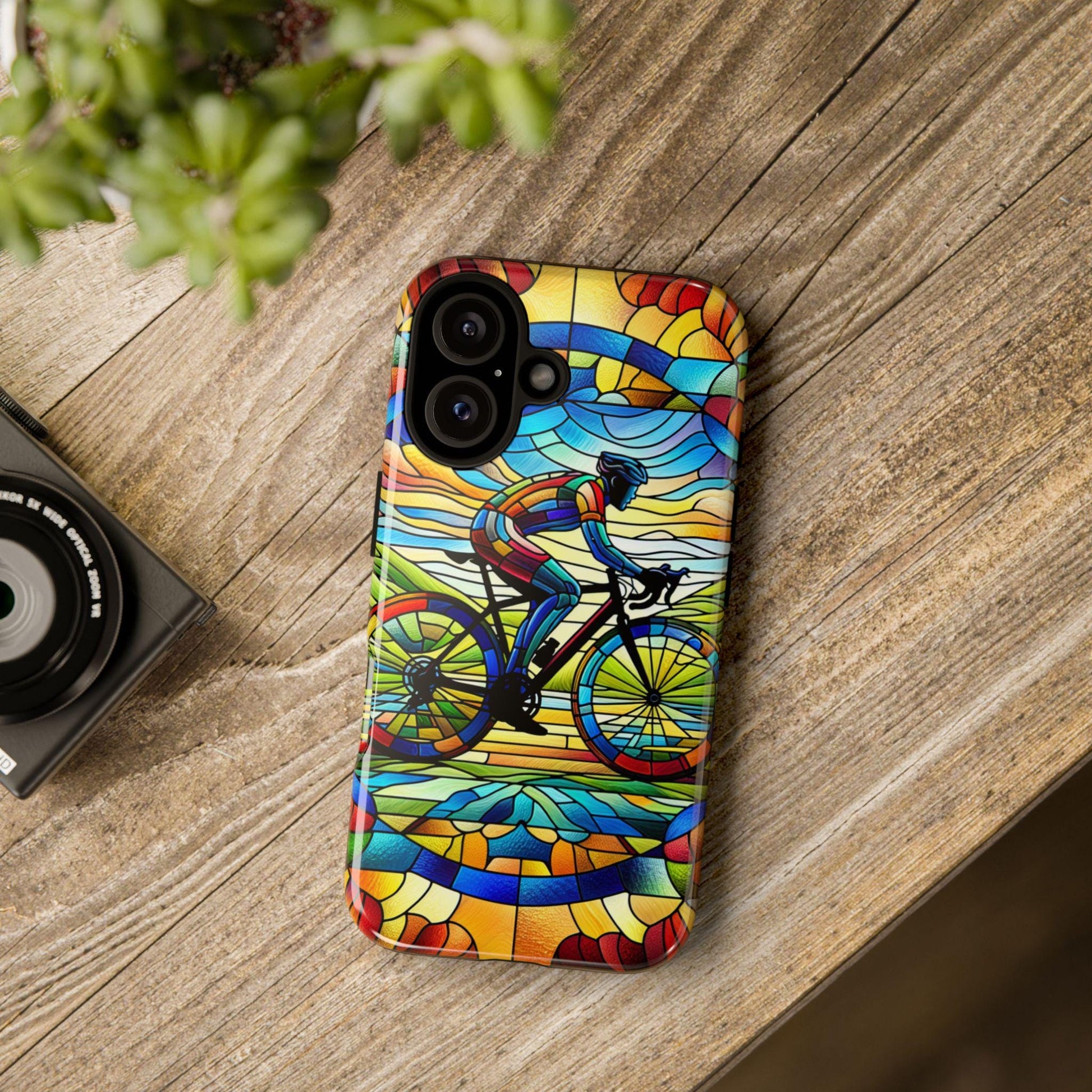 A smartphone with a Cycling Phone Case, Faux Stained Glass Cover For iPhone 16 15 14 13 Pro Samsung Pixel, featuring a cyclist design, lies on a wooden table beside a camera and potted plant.