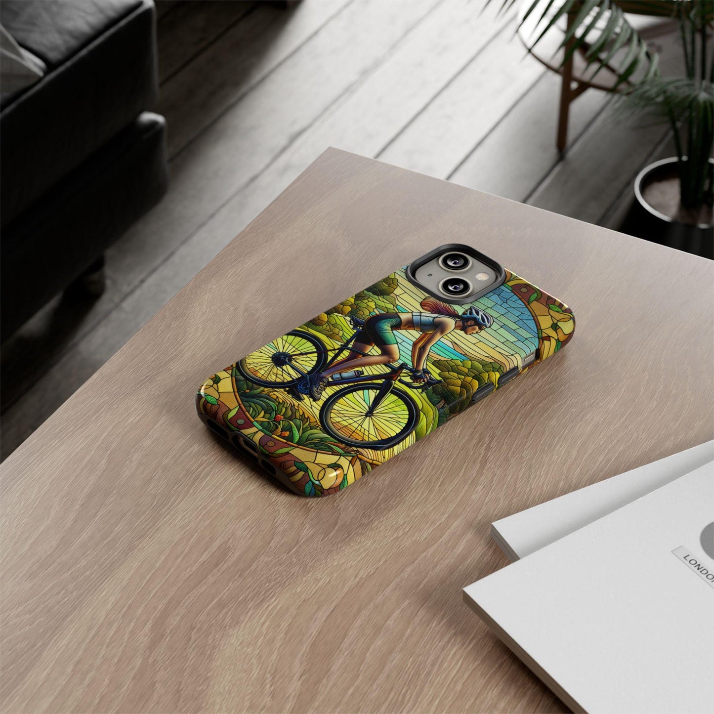 On a wooden table sits a phone adorned with the Female Cyclist Cycling Phone Case, showcasing a cyclist navigating a scenic path in a faux stained glass design.