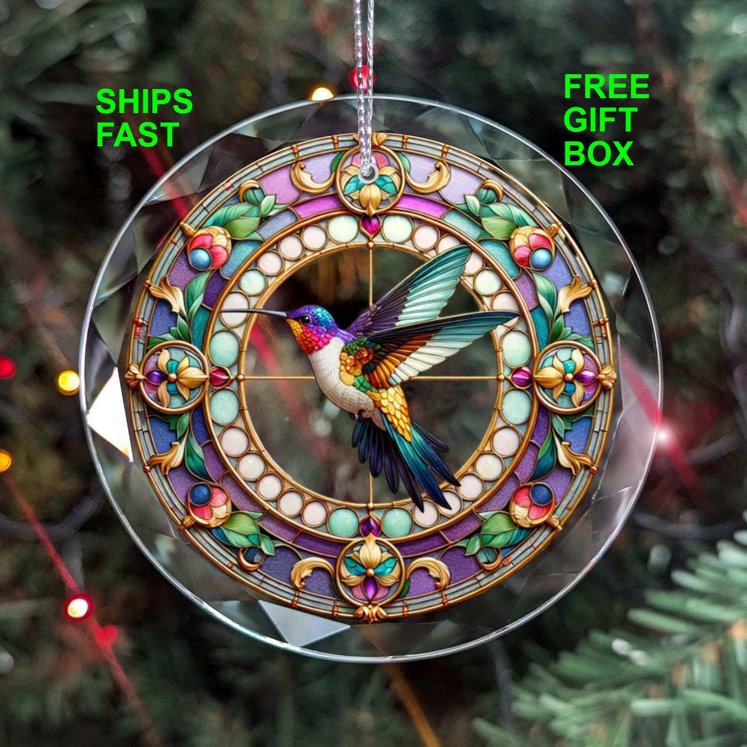 A Hummingbird GLASS Ornament by Golden Value SG, showcasing a colorful hummingbird in the center encircled by intricate floral patterns, decorates a Christmas tree, with blurred lights providing a festive backdrop.