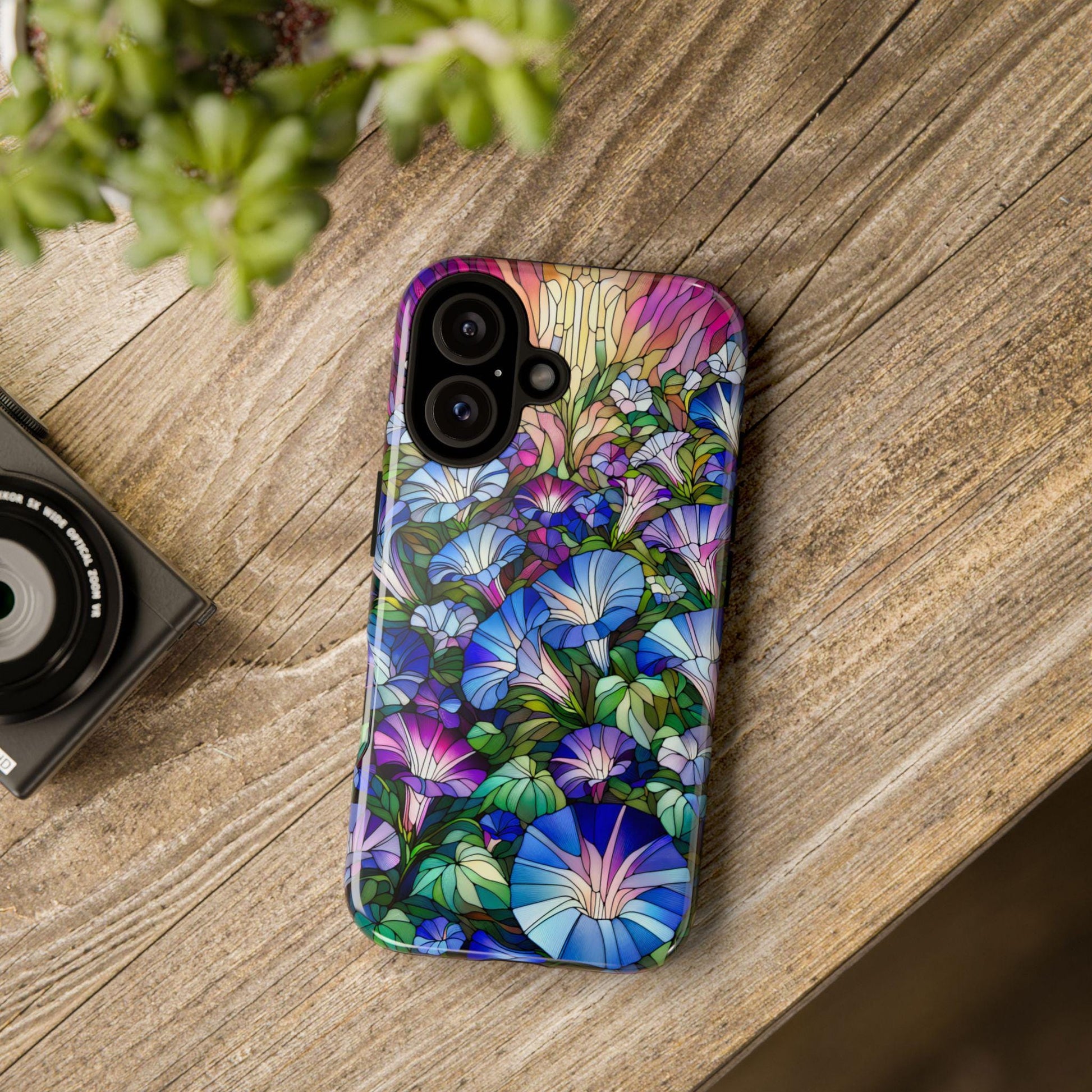 A smartphone with a September Birth Flower Morning Glory Phone Cover, featuring a faux stained glass wildflower design, rests on a wooden table beside a camera and a plant.