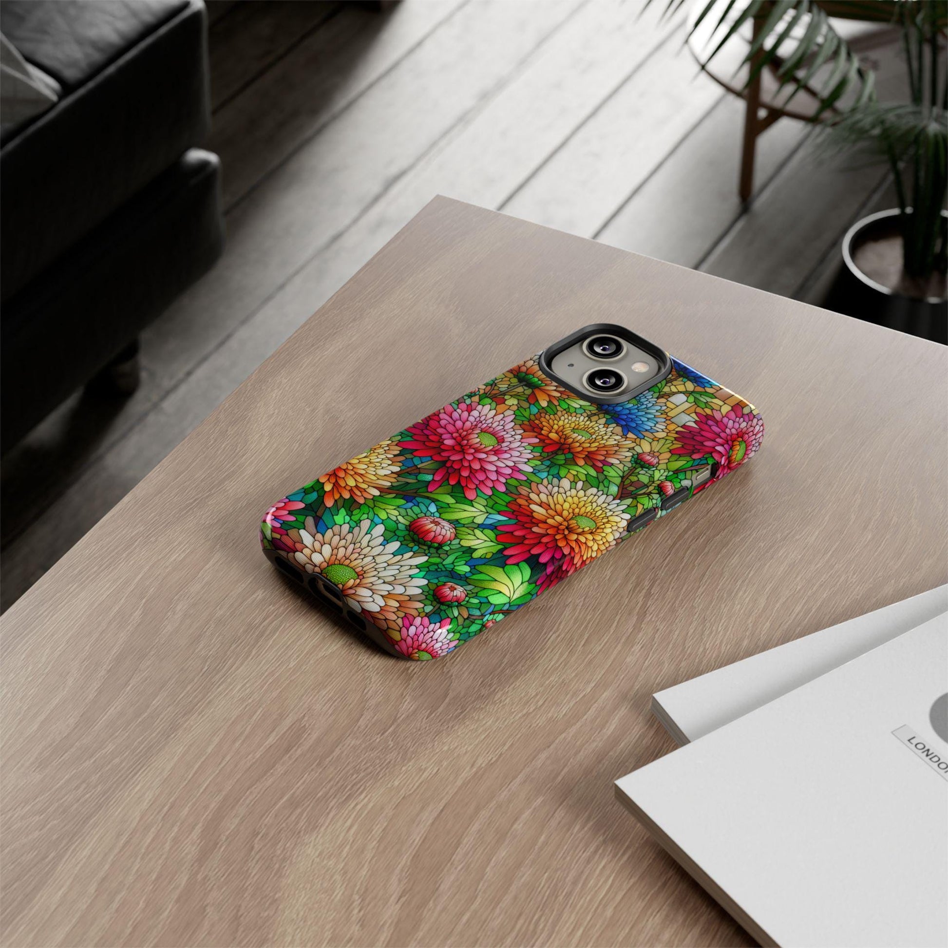 An iPhone with a November Birth Flower Chrysanthemum phone case, featuring a faux stained glass design, sits on a wooden table beside two closed books.