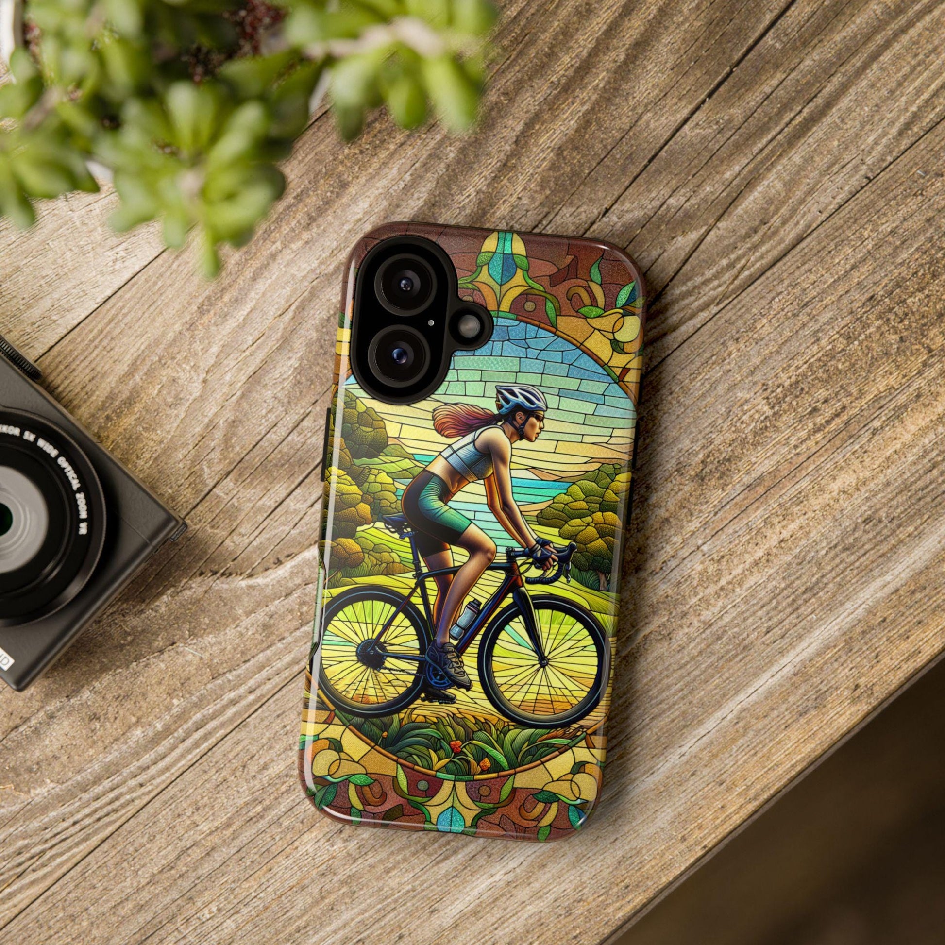 On a wooden table rests a Female Cyclist Cycling Phone Case with a faux stained glass nature design. A camera and plant nearby complete the scene, making it an ideal birthday or Xmas gift for any bicycle lover. Compatible with iPhone 16-13 Pro, Samsung, and Pixel models.