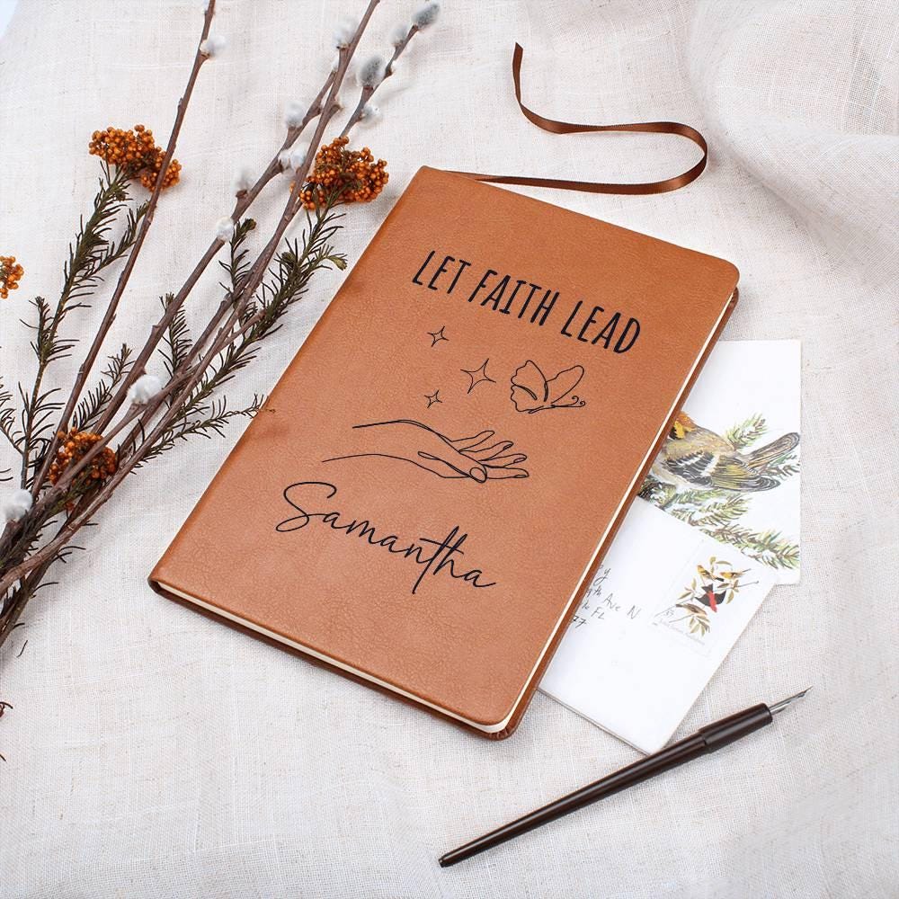 The Personalized Leather Prayer Journal for Women, Let Faith Lead Custom Name Christian Faith Gift Devotional Notebook with Ribbon Bookmark, and pen rest gracefully on a white cloth, inviting reflection.