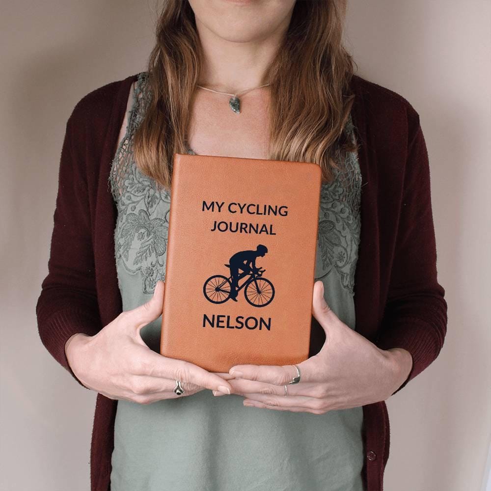 A woman in a maroon cardigan and green shirt holds a Personalized Leather Cycling Journal For Men, Custom Name Notebook with “MY CYCLING JOURNAL NELSON” and a cyclist illustration on the cover. Only her torso and hands are visible.