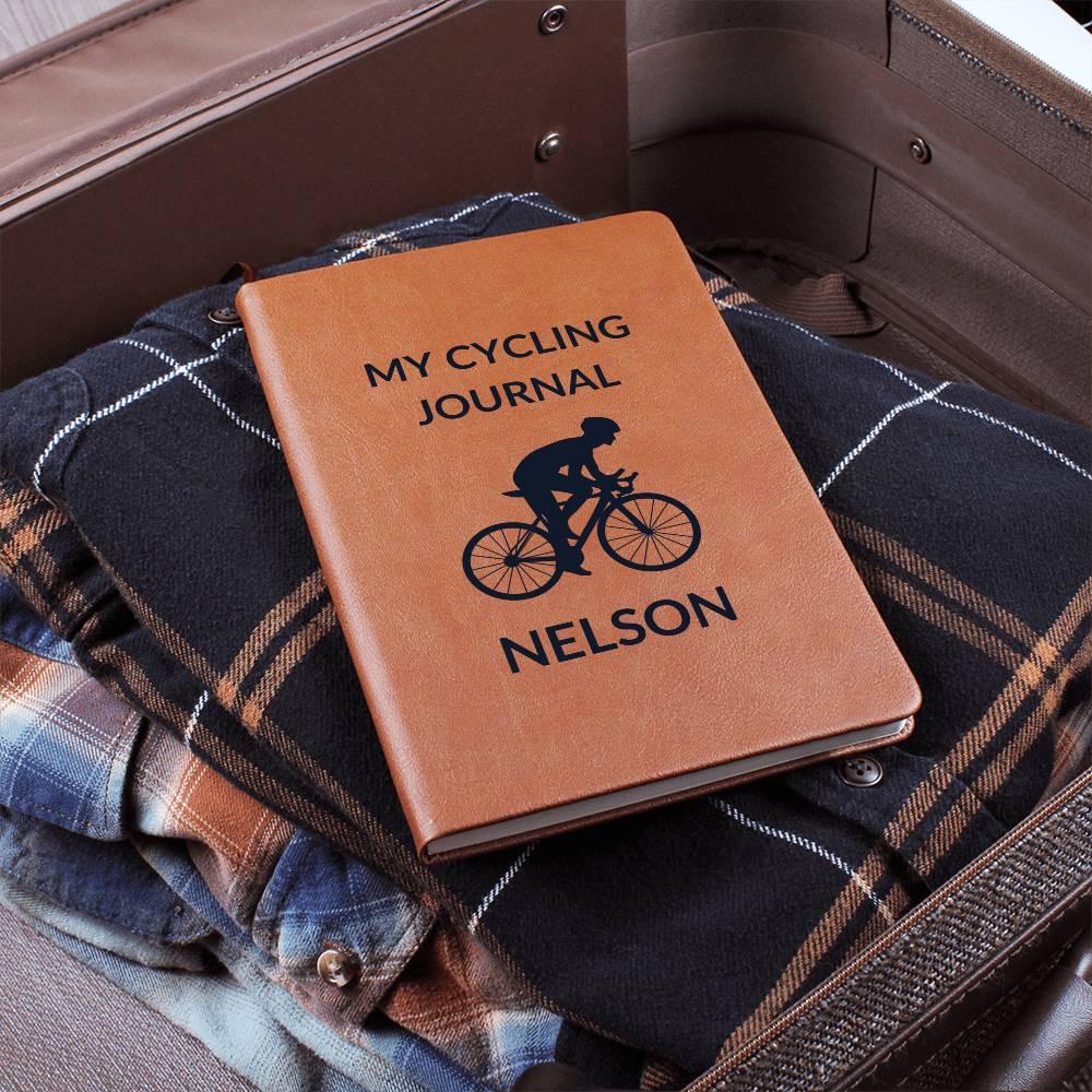 A personalized leather cycling journal for men titled "My Cycling Journal" with "Nelson," featuring a cyclist's silhouette, rests on plaid fabric in an open suitcase—a perfect companion for tracking every mile.