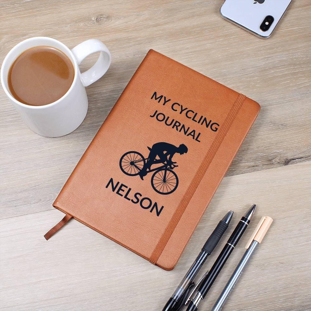 A personalized leather cycling journal labeled "My Cycling Journal Nelson" sits on a table with coffee, a smartphone, and three pens. This brown notebook is perfect for road bikers and bike lovers, featuring a ribbon bookmark.